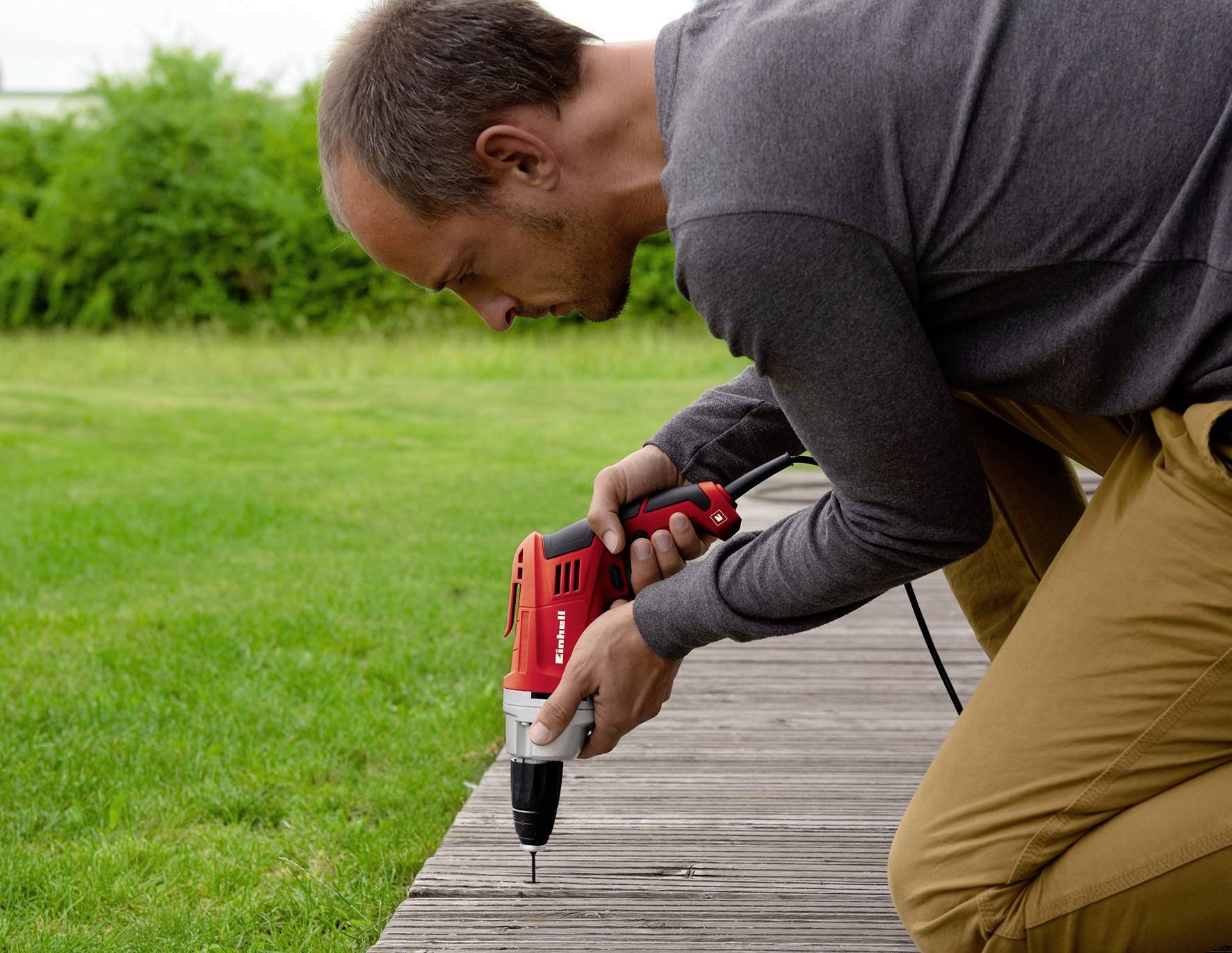 Egy férfi térdel, és egy piros fúrógéppel lyukakat fúr a fadeszkákból készült teraszon, miközben zöld fű veszi körül.