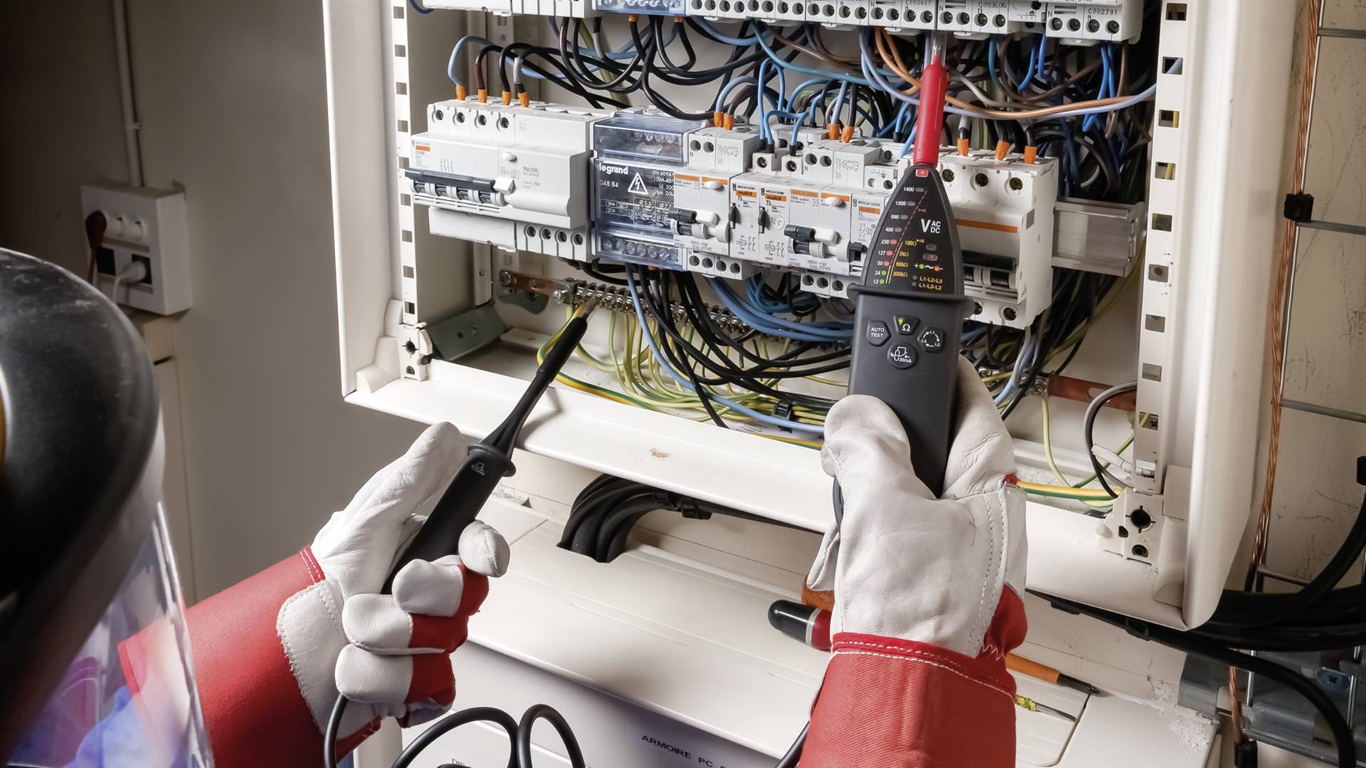 Villanyszerelő védőfelszerelésben ellenőrzi az elektromos kapcsolóberendezést műszerek segítségével.