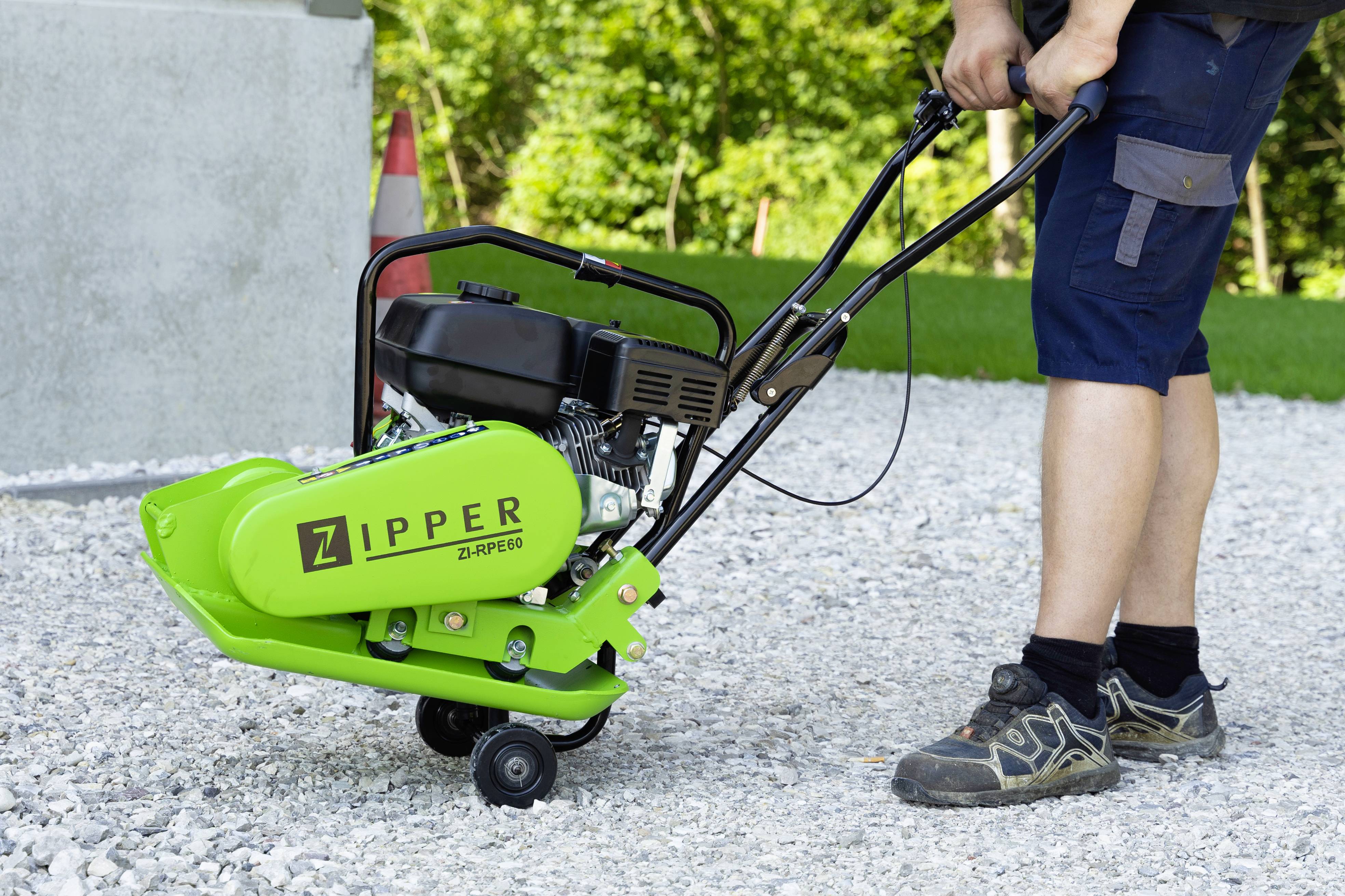 Egy személy egy zöld Zipper márkájú tömörítő lapot kezel egy kavicsos talajon a szabadban.