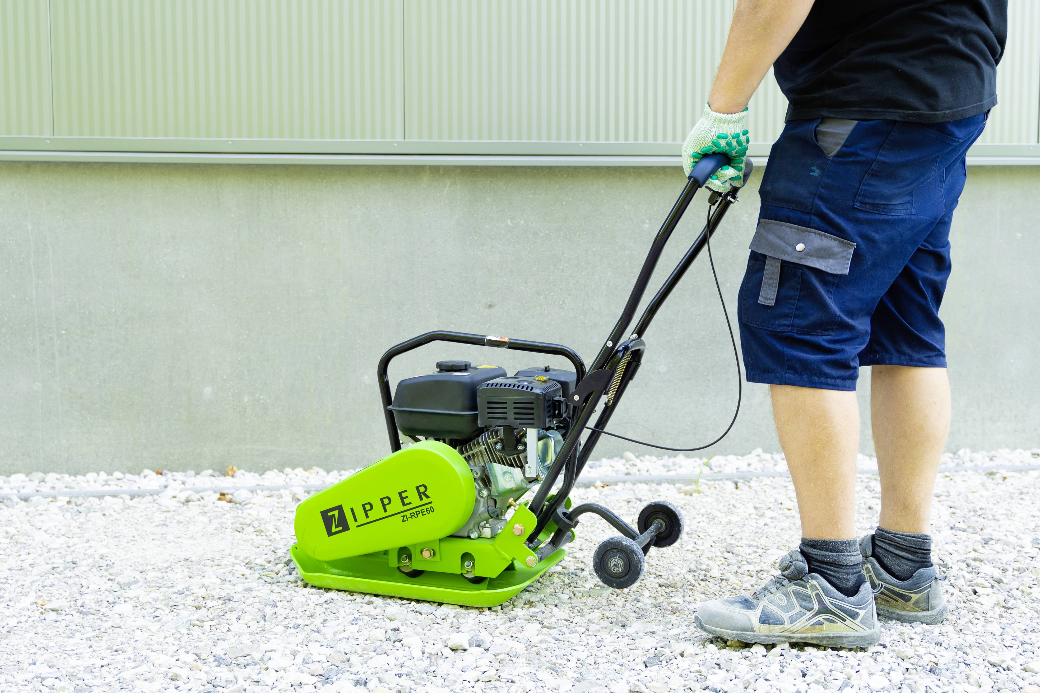 Egy ember egy zöld, 'Zipper' márkájú tömörítőlappal zúzalékot tömörít egy szürke fal előtti alapfelületen.