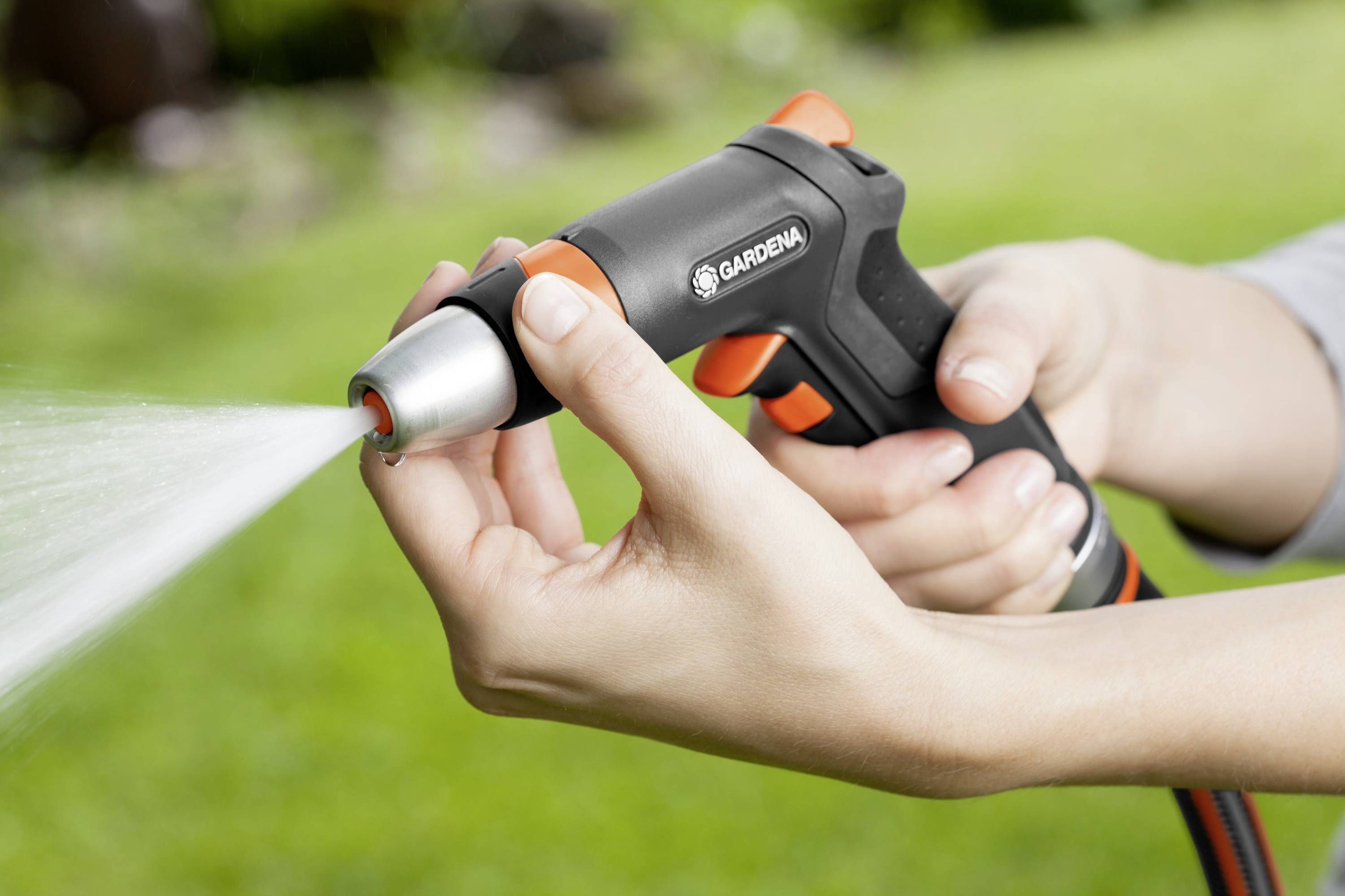 Egy ember egy fekete és narancssárga kerti tömlőfejjel locsol, amelyből víz spriccel egy zöld, elmosódott háttér előtt.