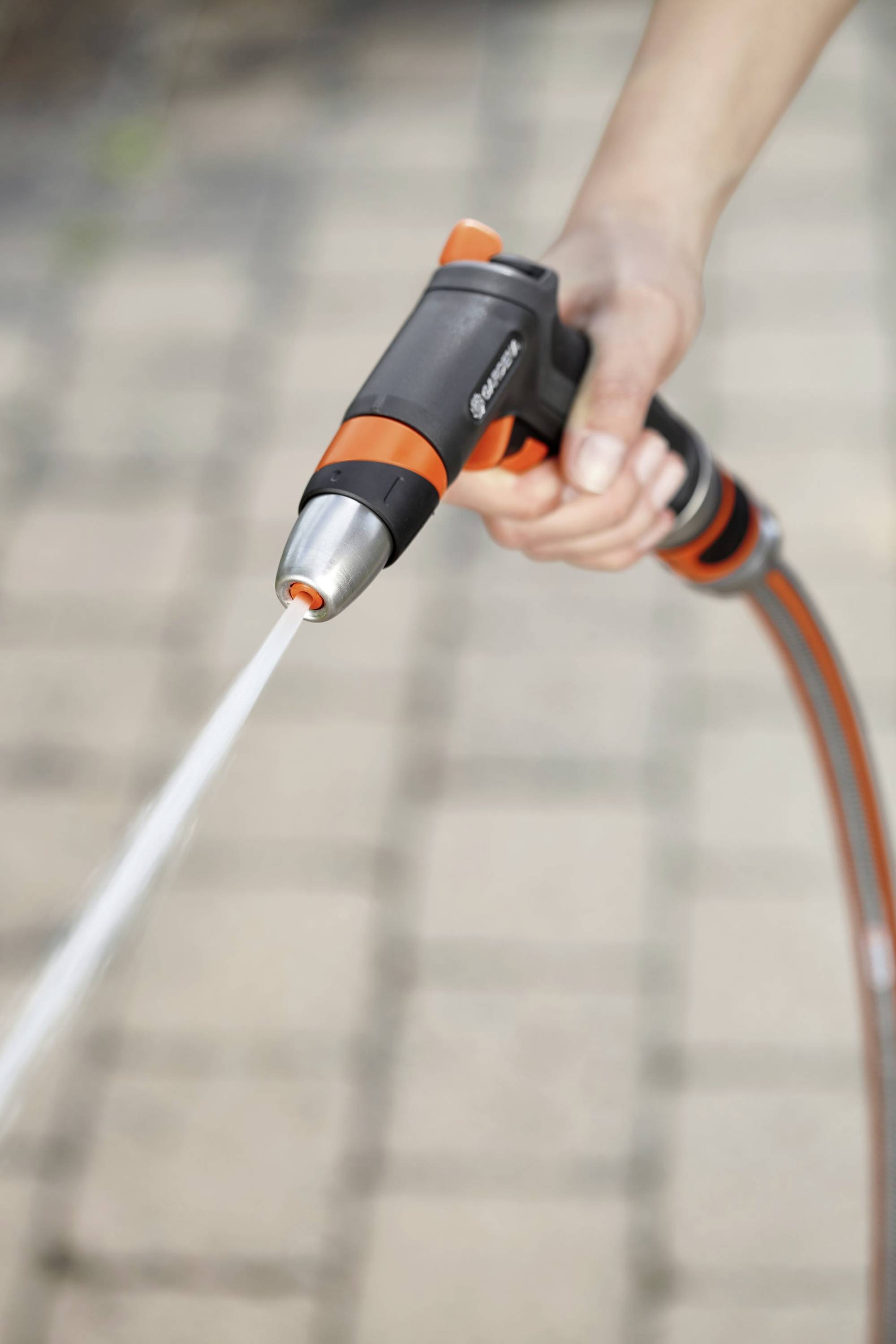 Egy kéz kerti tömlőt tart permetező fúvókával, amelyből vízsugár spriccel. A háttérben térburkolat látható.