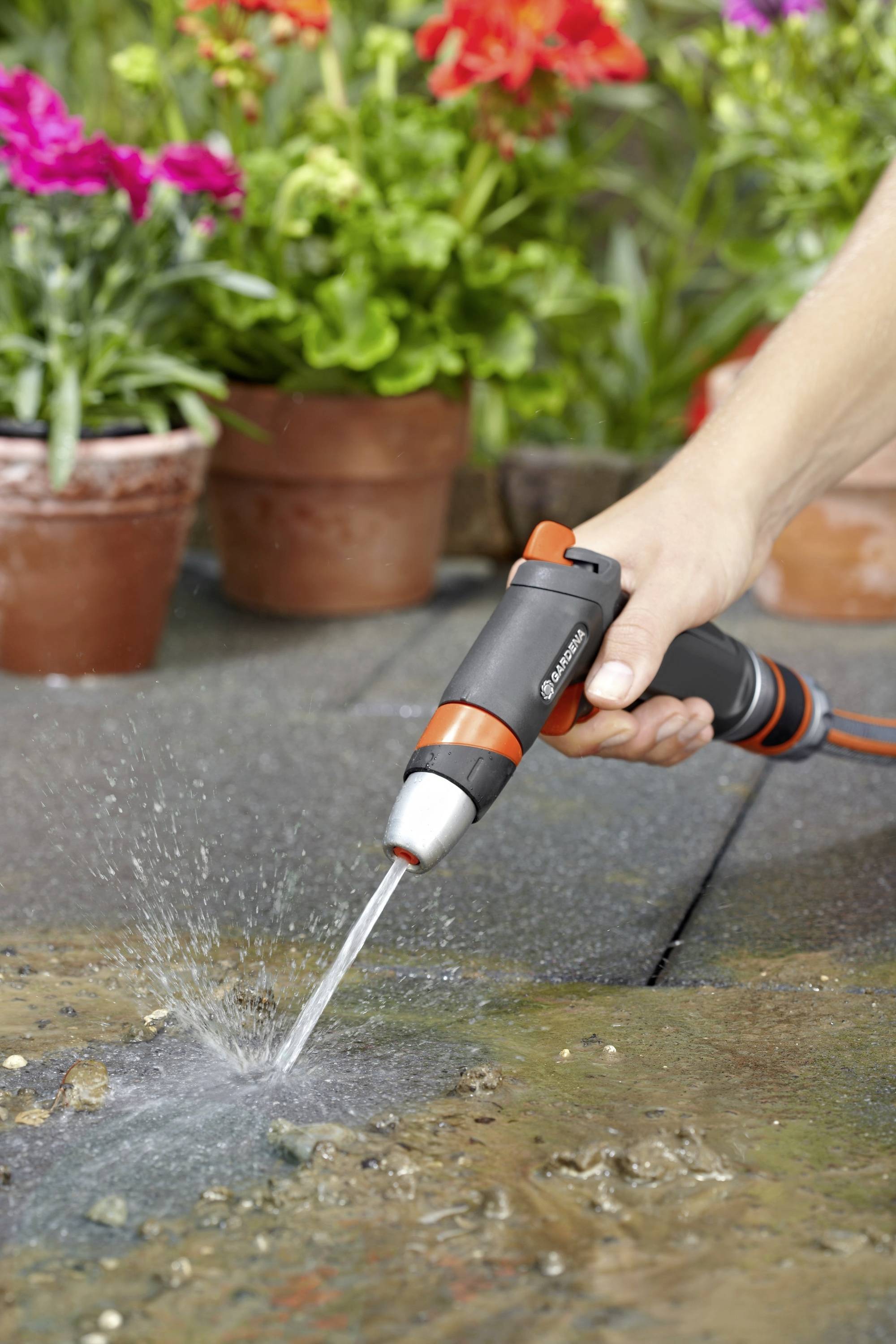 Egy ember egy kerti locsolóval öntözi a teraszát. A háttérben virágcserepek láthatók tarka virágokkal.