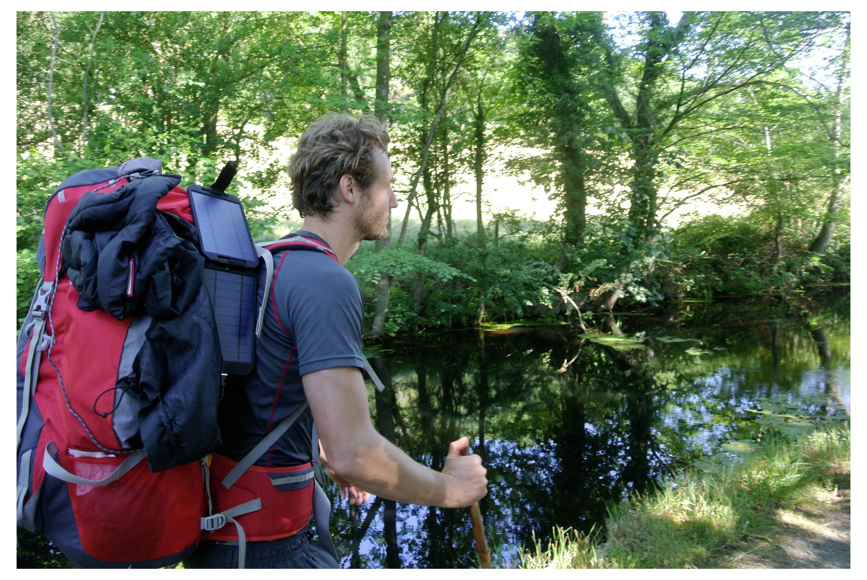 Egy nagy hátizsákos ember áll egy tükröződő erdei tó mellett, kezében egy túrabottal, dús zöld fák között.