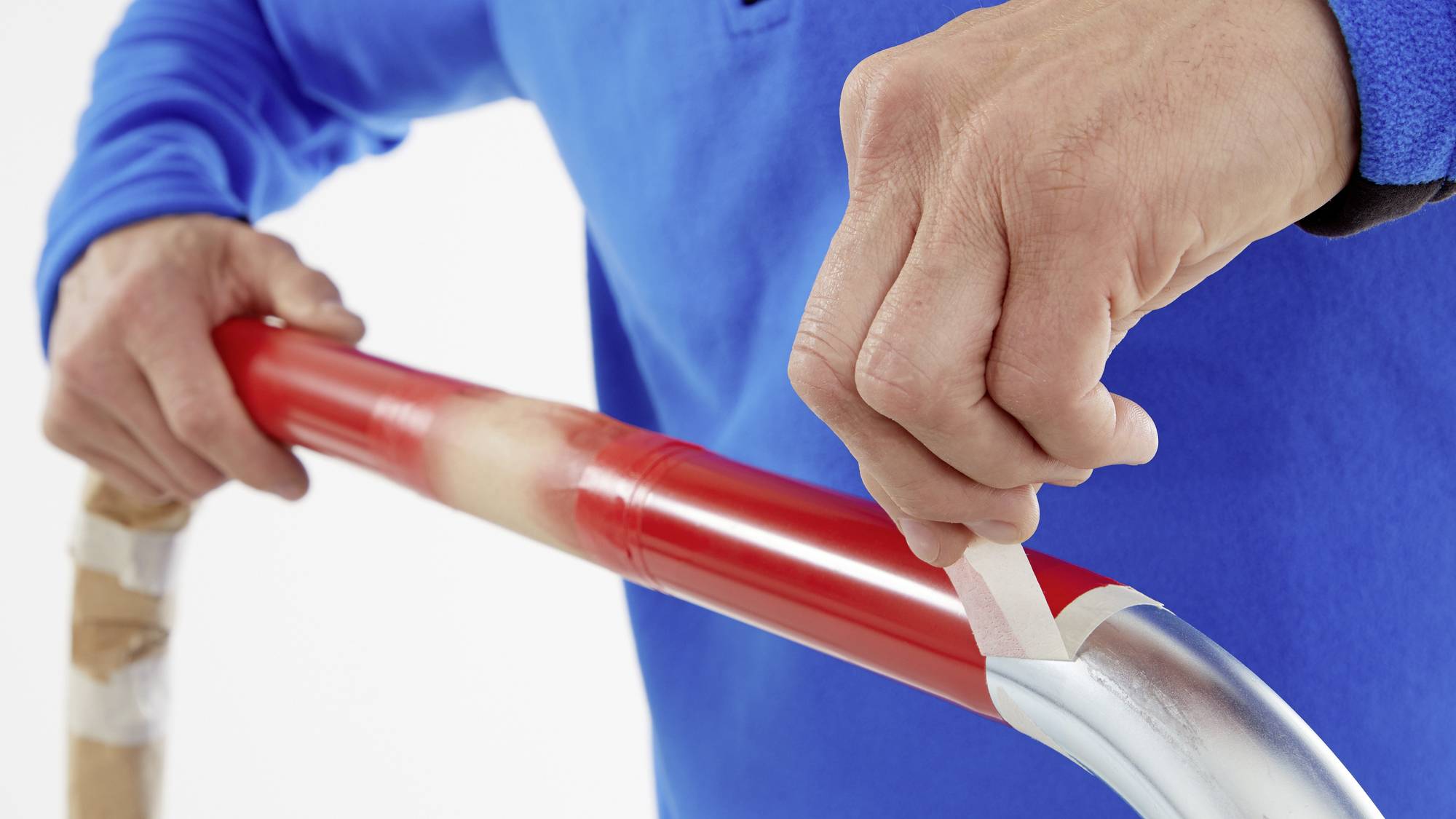 Egy kék pulóveres személy egy piros hokiütő nyelet fehér szalaggal ragaszt le. A kezek a precíz tekercselésre összpontosítanak.