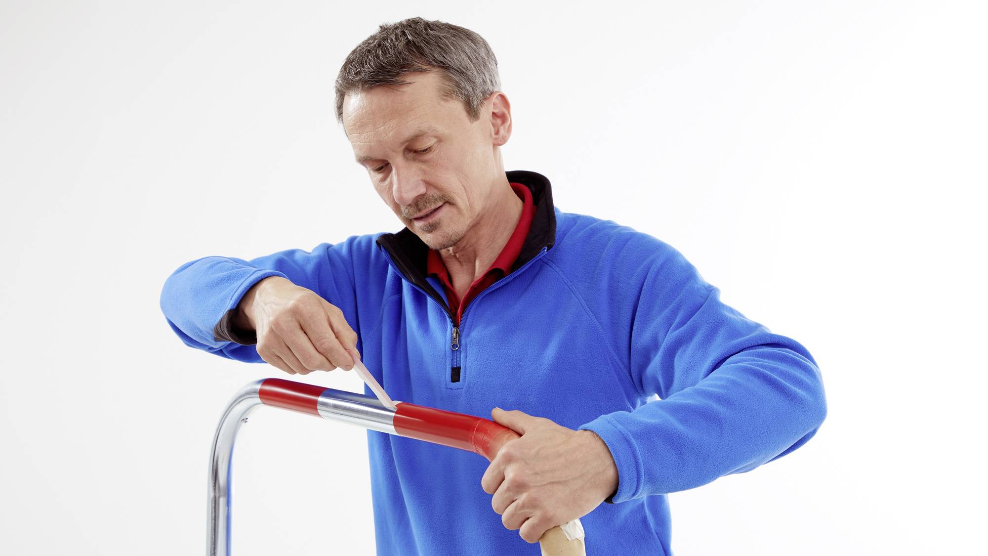 Egy férfi kék pulóverben egy szikével vág le egy piros szigetelőszalag végét egy fémrúdnál.