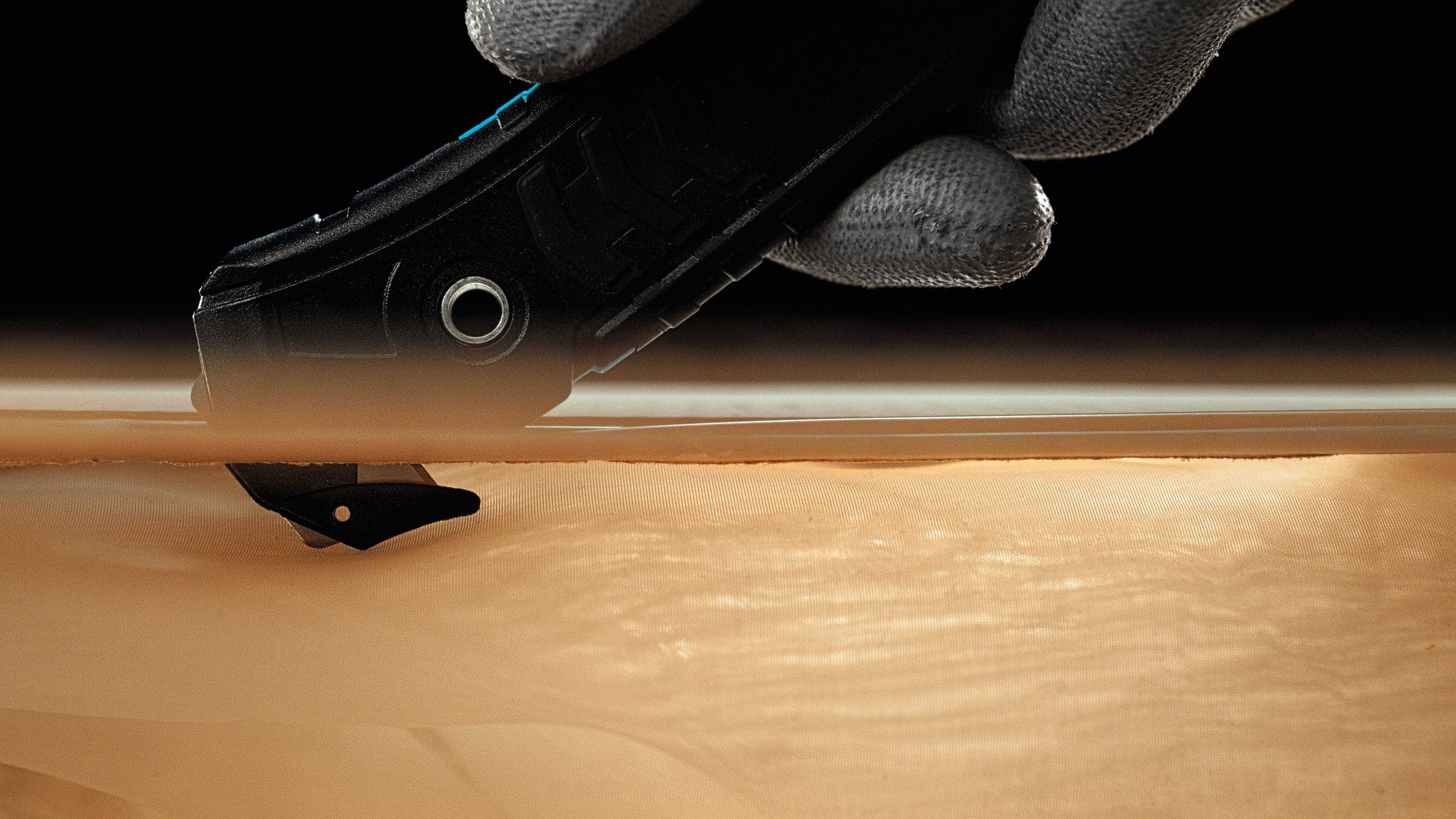 Egy szürke kesztyűs kéz egy fekete pengés vágókéssel egy sima, matt felületet vág.