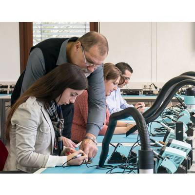 Egy tanár segít egy diáklánynak egy forrasztási gyakorlaton egy elektronikai laboratóriumban, miközben más diákok összpontosítva dolgoznak a saját projektjeiken.