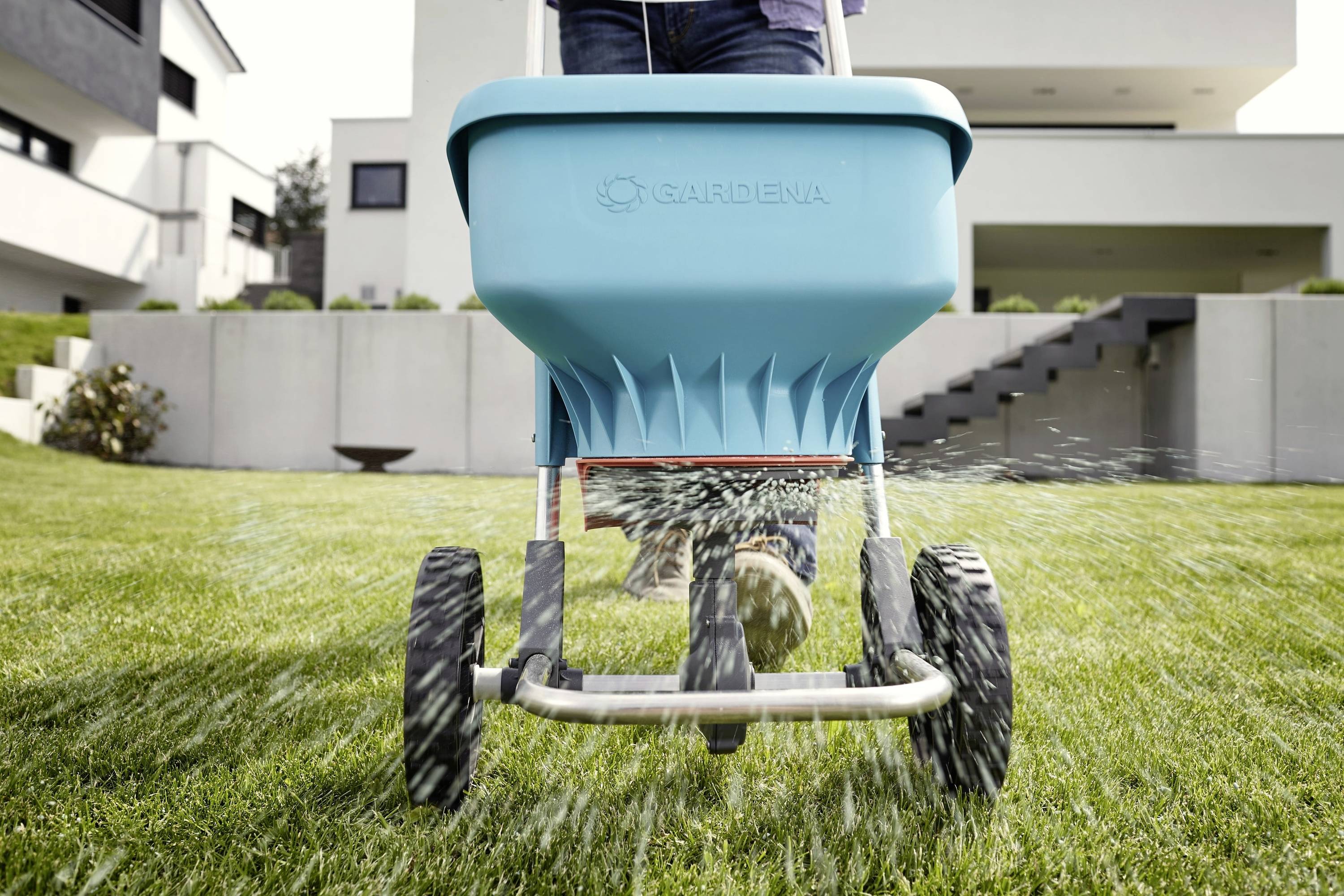 Egy személy egyenletesen szórja a fűmagot egy szórókocsival egy gondozott gyepfelületen egy modern ház előtt.