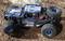 Egy 'Raptor 6S' és 'Arrma' feliratú kék és fekete RC-buggy áll a talajon. Az autónak nagy, durva mintázatú kerekei vannak.