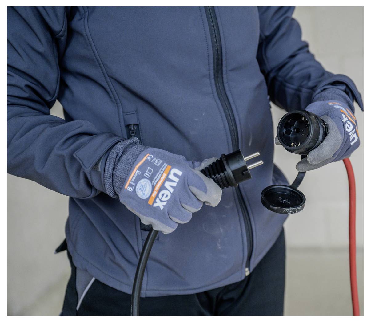 Kesztyűben és dzsekiben lévő személy egy elektromos dugót és csatlakozót vizsgál, egy piros kábel csatlakoztatva.