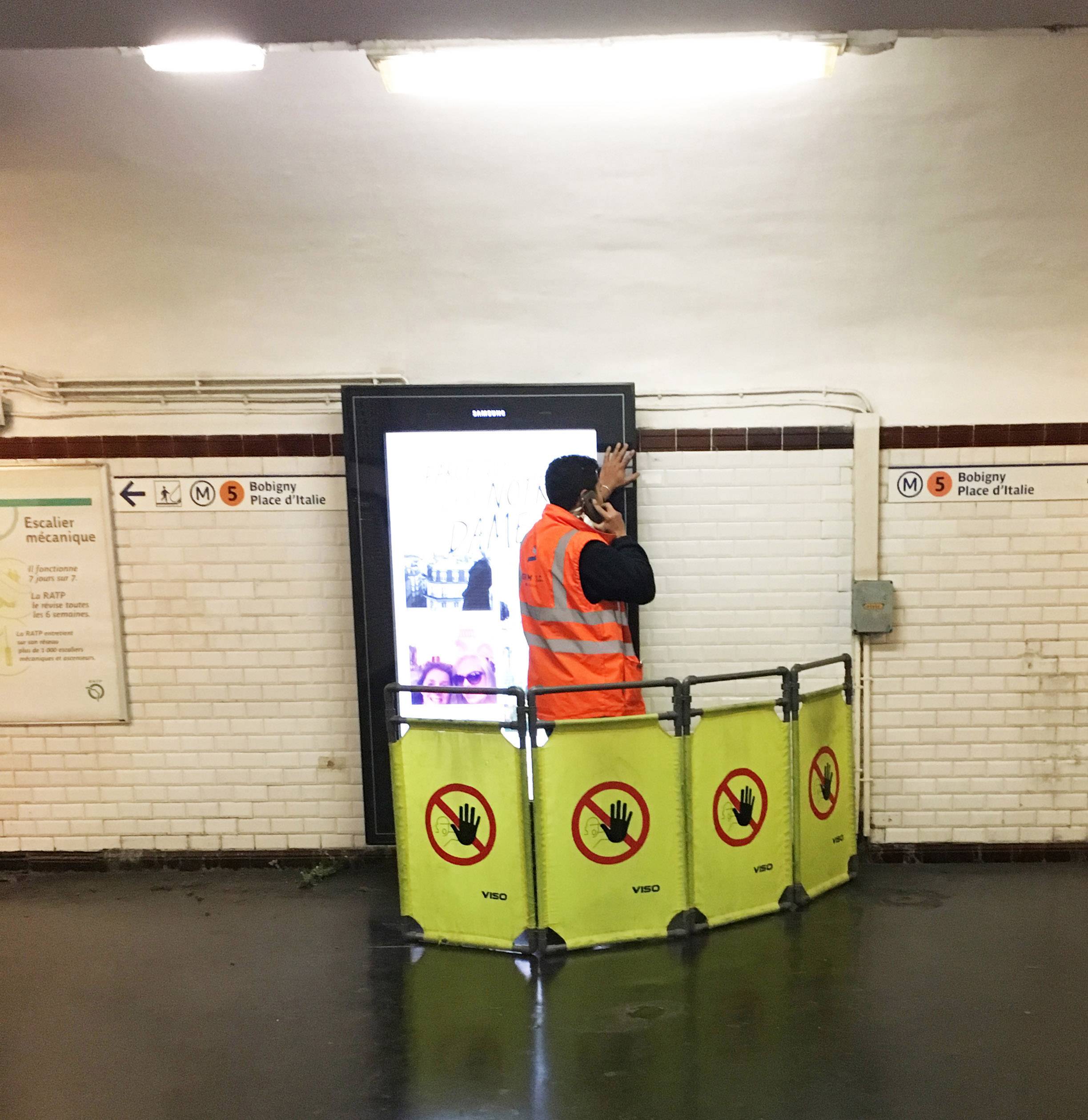 Egy narancssárga figyelmeztető mellényt viselő munkás egy hirdetőtáblát töröl egy metróállomáson. Körülötte védőkorlátok vannak.