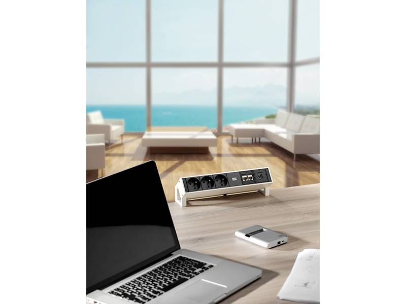Egy laptop, powerbank és elosztó egy fa asztalon egy modern nappali térben, nagyméretű ablakokon át kékesen csillogó óceáni kilátással.