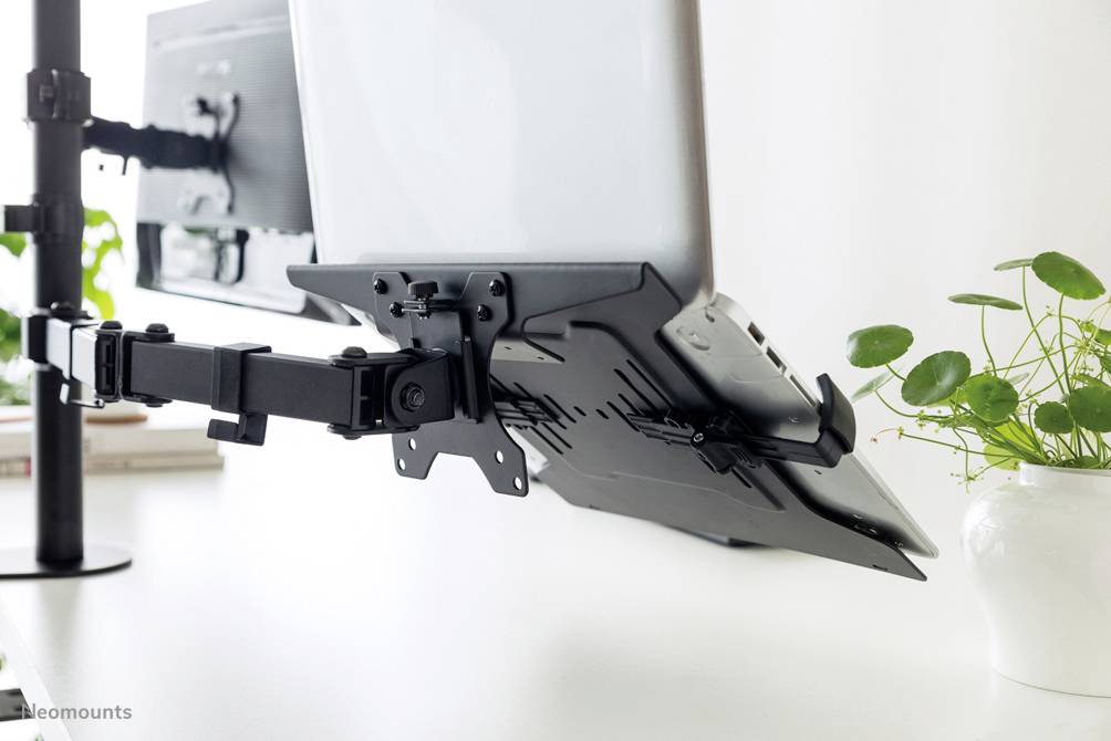 Laptop-tartó egy állítható karral, amely egy íróasztalhoz van rögzítve, növényekkel a háttérben. Ideális ergonomikus munkaterületekhez.