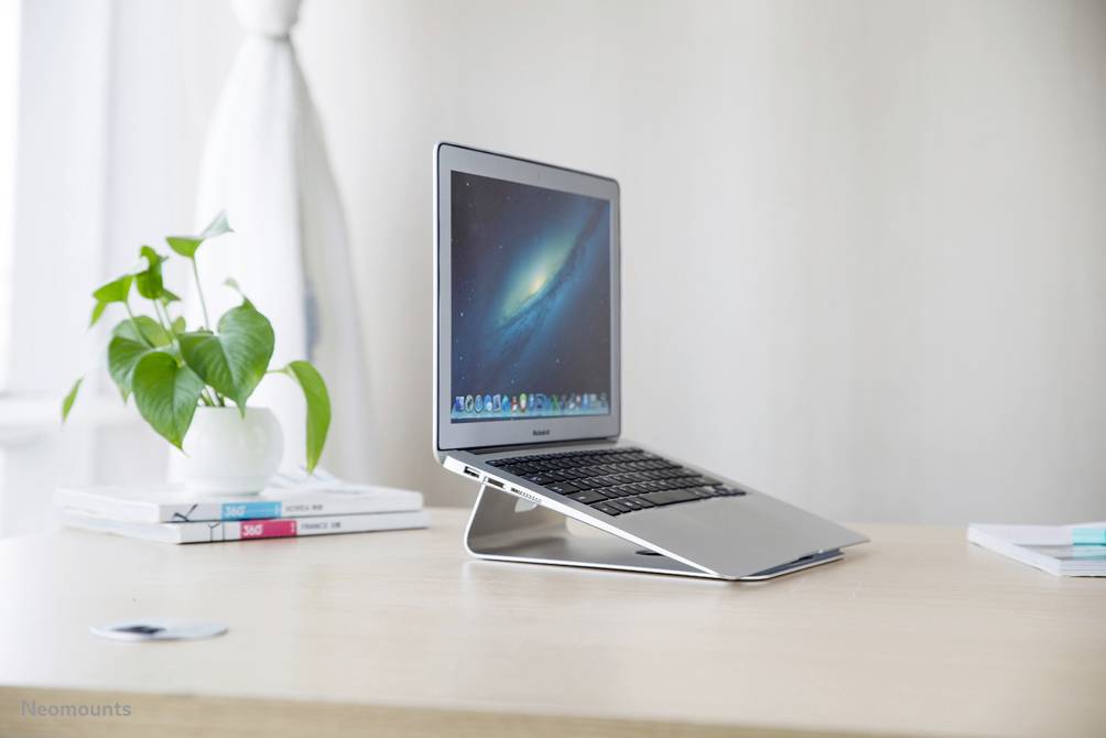 Egy laptop áll egy asztalon egy tartóval, egy zöld növényt tartalmazó cserép mellett. A képernyőn egy világűr háttérkép látható.