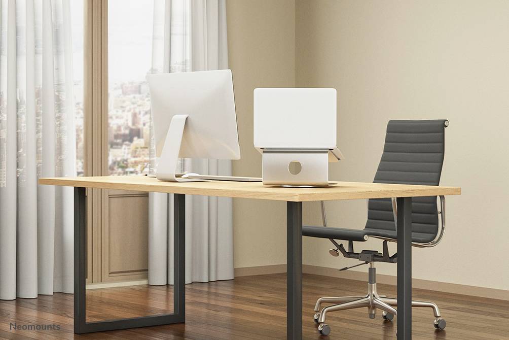 Modern íróasztal számítógéppel és ergonomikus székkel egy világos irodában, ablak mellett.