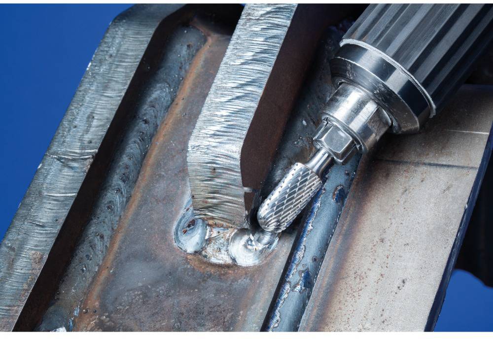 Egy fémmunkás közeli felvétele, amint egy marófréssel hegesztési varratokat simít el. Látható fémréteg és kék háttér.