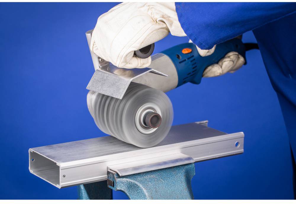 Egy személy egy kék elektromos csiszológéppel csiszol egy fémdarabot. Fehér védőkesztyűt visel.
