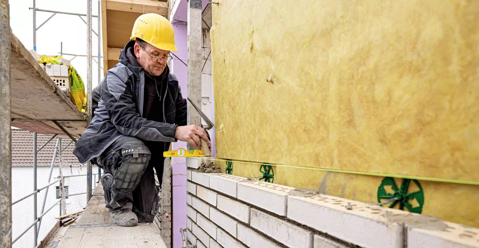 Egy sárga védősisakot viselő építőmunkás egy vízmértékkel igazítja a téglák egyenességét egy szigetelt falon, állványzaton állva.
