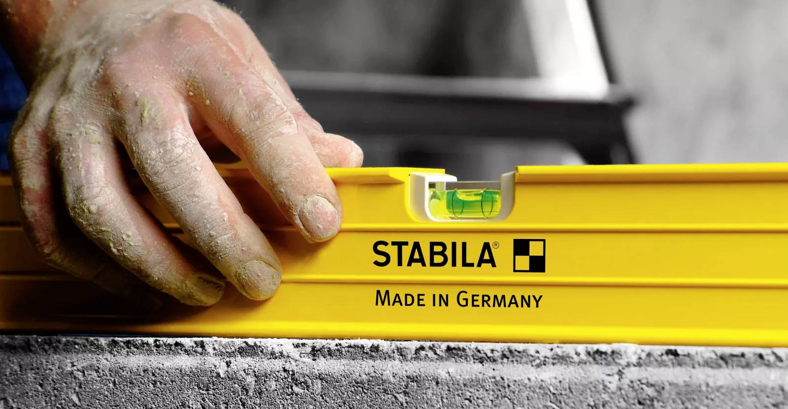 Egy kéz egy sárga vízmértéket állít be, amelyen a 'STABILA Made in Germany' felirat szerepel, egy szürke betonfelületen, gondoskodva arról, hogy tökéletesen vízszintes legyen.