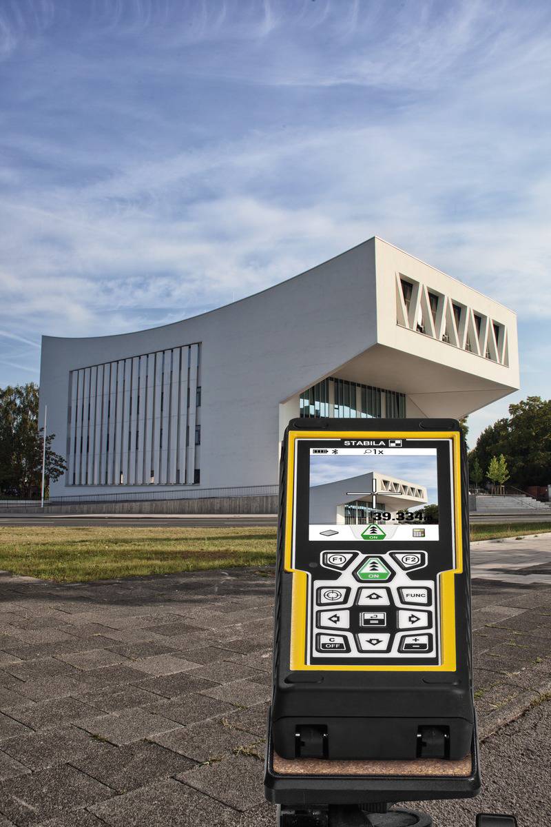 Egy fehér, modern épület ferde tetővel áll a háttérben, míg egy lézeres távolságmérő látható az előtérben.