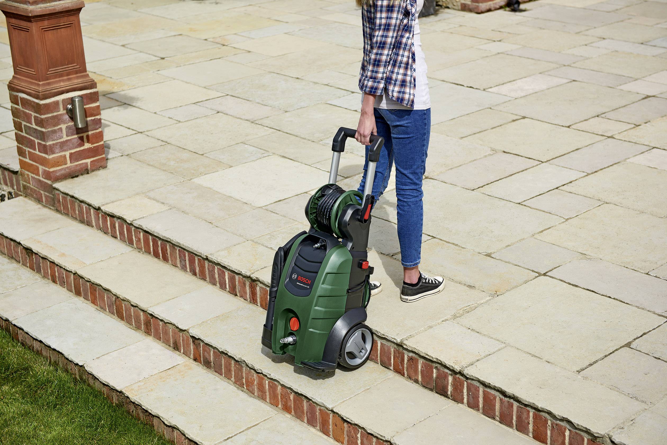 Egy ember egy magasnyomású mosót húz kőlépcsőkön. A gép kerekekkel és tömlővel rendelkezik, szabadtéri használatra alkalmas.