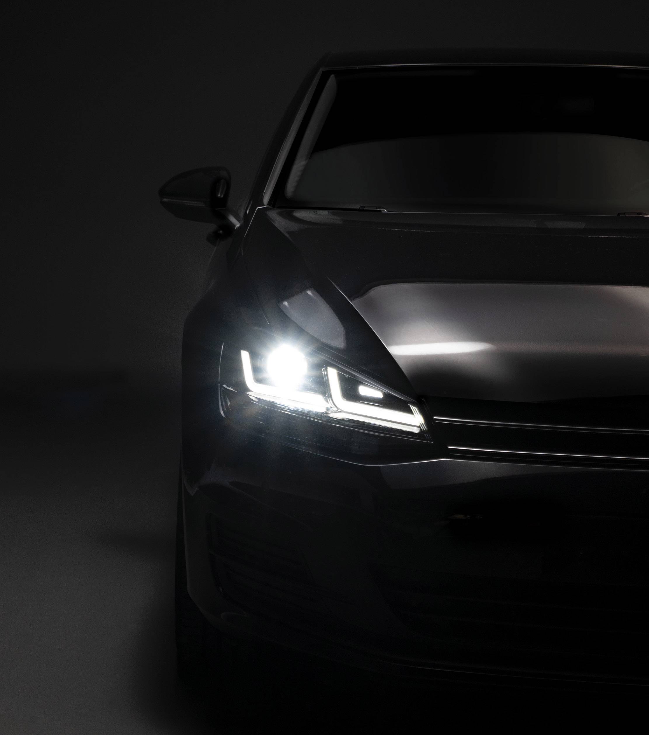 Sötét autó bekapcsolt fényszóróval, oldalról látható, sötét háttér előtt kiemelve.