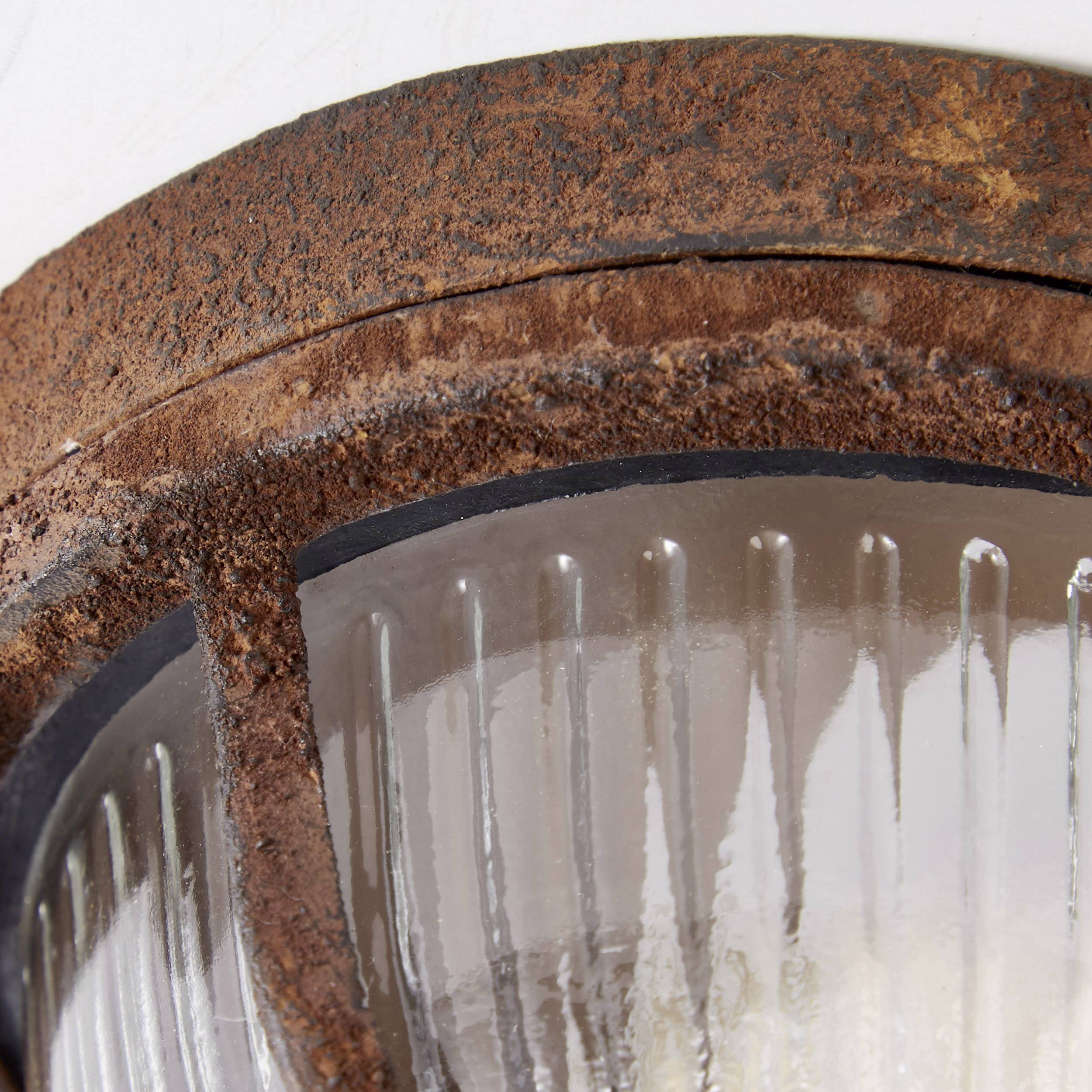 Egy rozsdás fémlámpáról közeli felvétel egy falon. A lámpa be van kapcsolva, és fénye átszűrődik a bordázott üvegen.