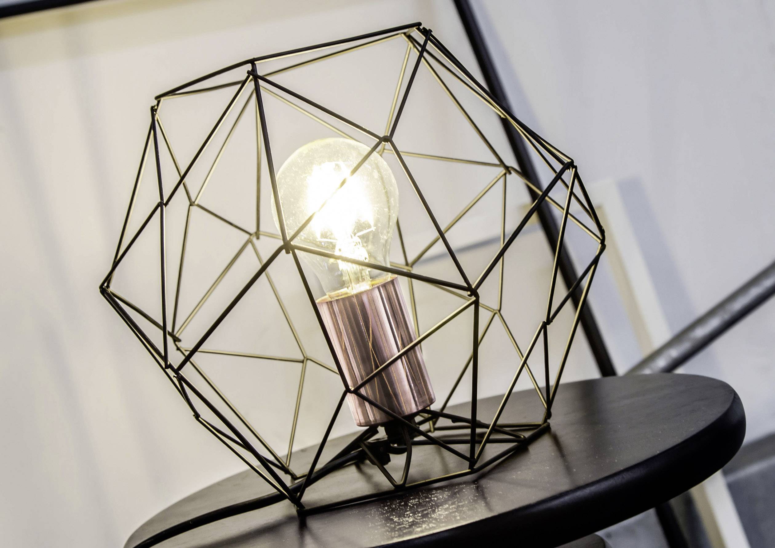 Egy villanykörte egy modern, geometrikus fémkeretben áll az asztalon. A lámpa világít, ami használatra utal.