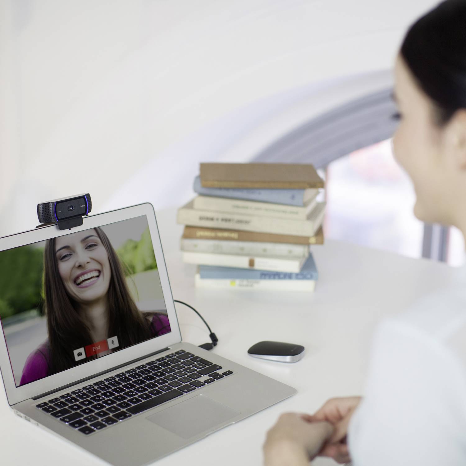 Egy személy videóhívást folytat egy laptoppal, mosolygó nő látható a képernyőn. A háttérben könyvek vannak egy asztalon egymásra pakolva.