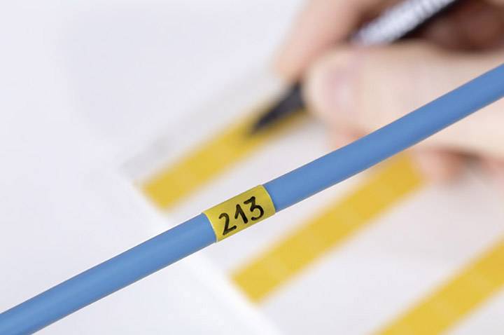 Egy kéz egy sárga diagramokkal teli papír fölött tart egy tollat. Az előtérben egy kék kábel a '213' számmal.