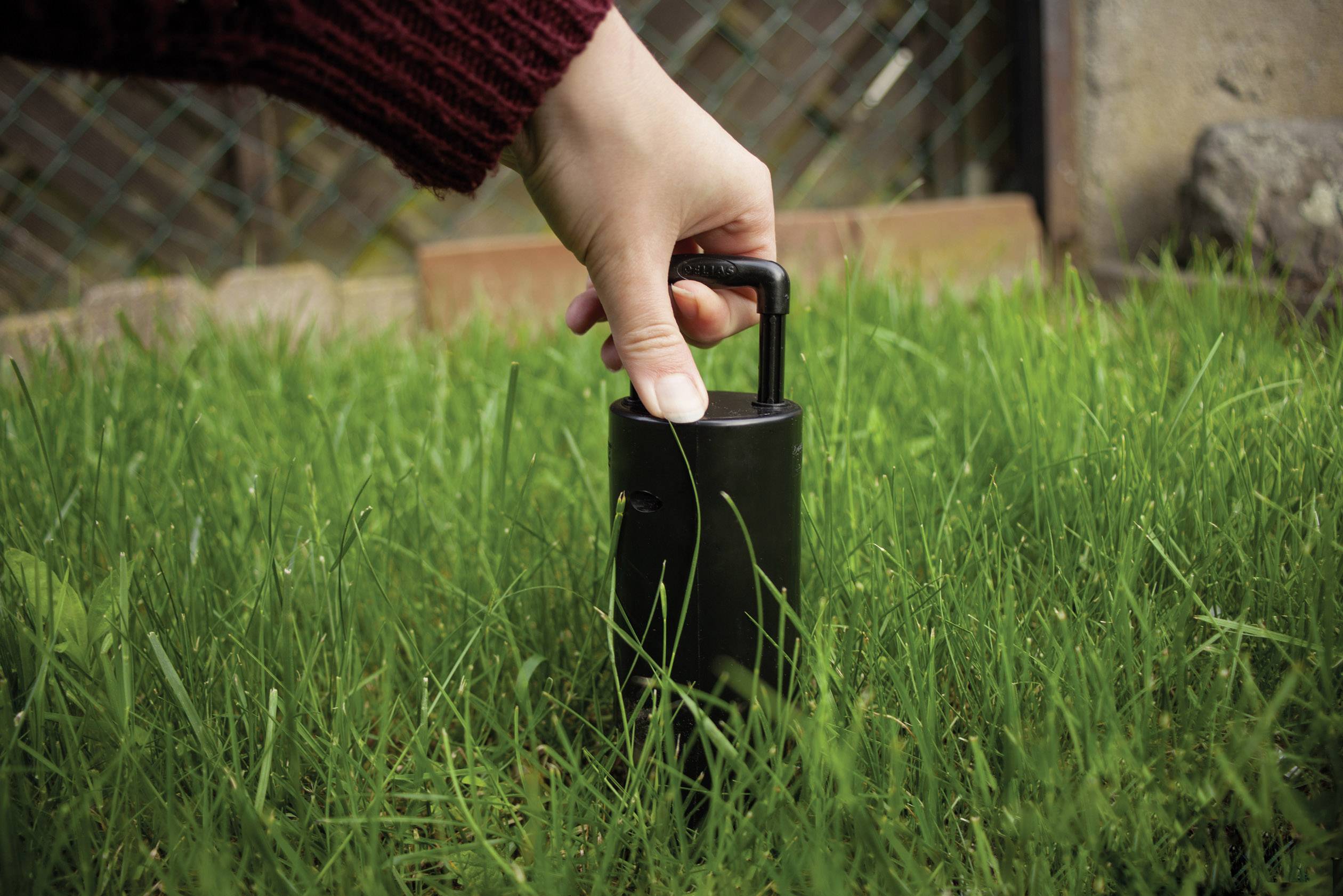 Egy kéz egy fekete hengert emel ki a fűből, valószínűleg egy kerti eszközt vagy egy kisebb készüléket egy kertben.