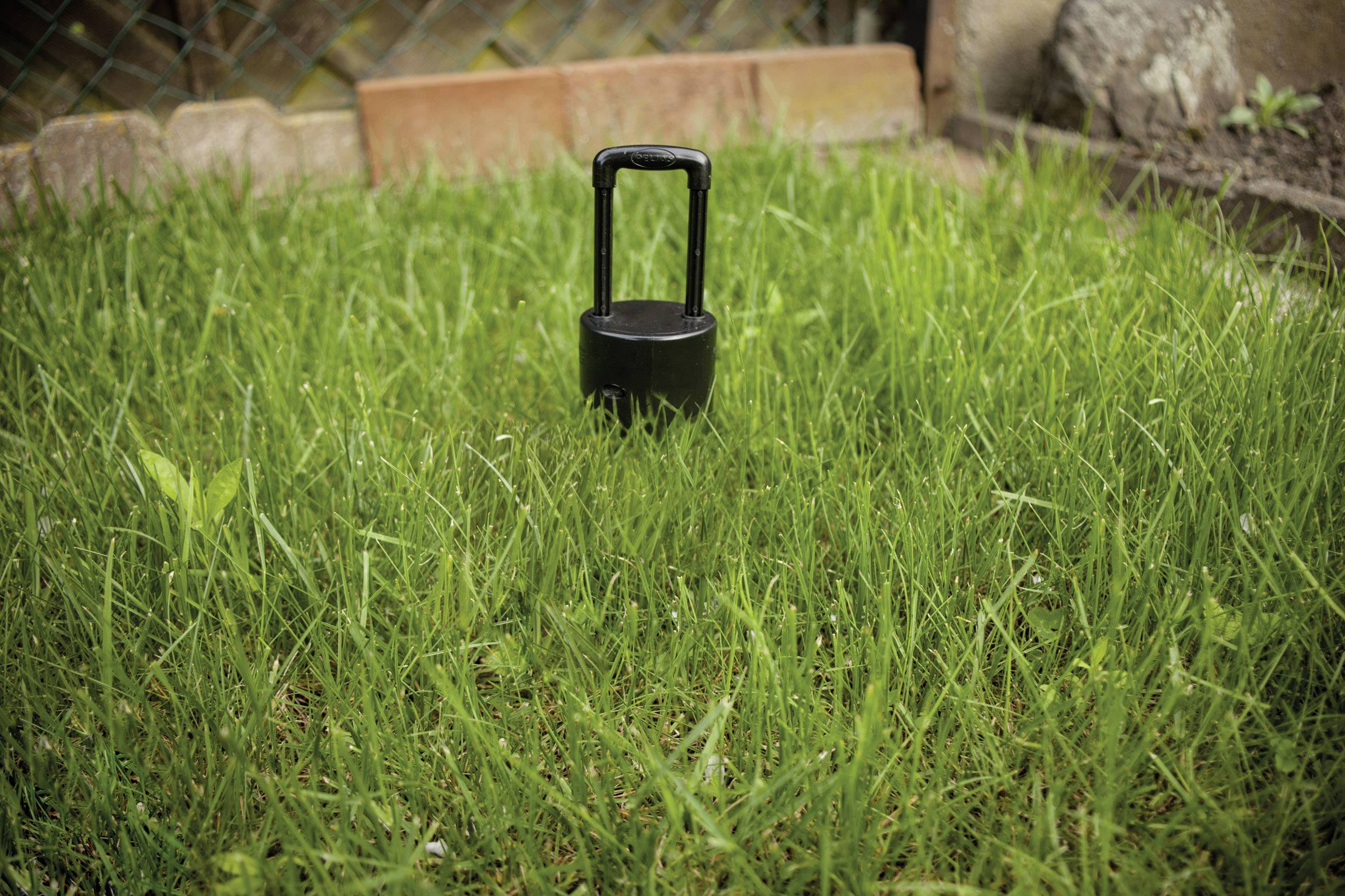 Egy fekete növényvédőszer-adagoló áll egy gyepfelületen egy kertben. A háttérben egy fakerítés látható.