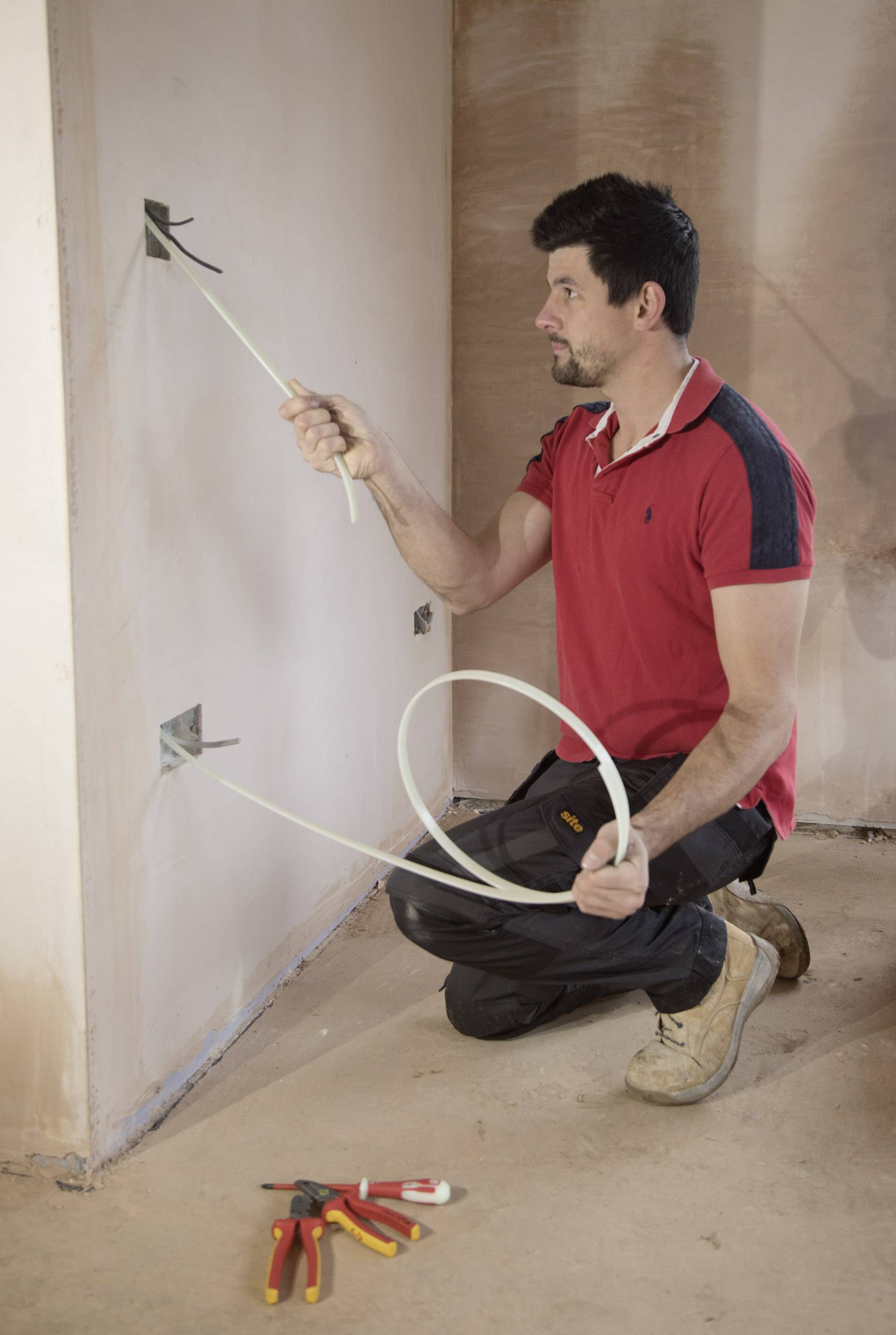 Egy férfi elektromos kábeleket szerel egy falba. Mellette szerszámok fekszenek a padlón. Piros inget és fekete nadrágot visel.