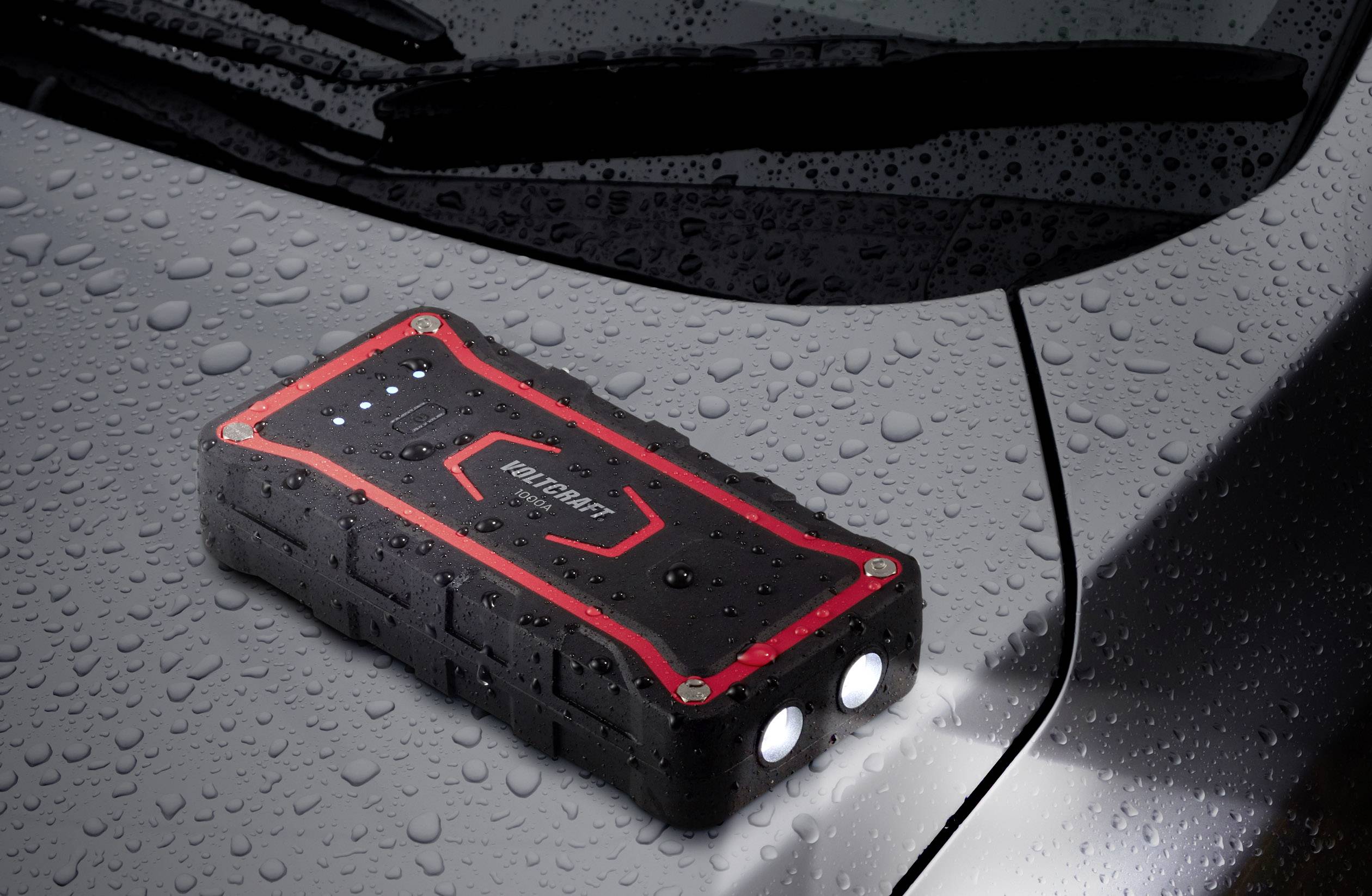 Egy fekete, hordozható autóakkumulátor-indító piros kiemelésekkel és LED-lámpákkal egy autó nedves motorháztetején fekszik.