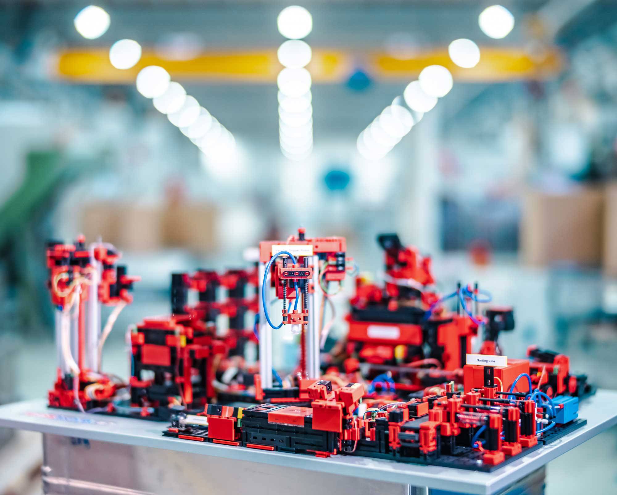 Egy miniatűr gyár vörös építőkockákból álló épülete komplex gépeket és robotkarokat mutat egy világosan megvilágított munkakörnyezetben.