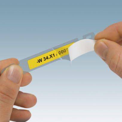 Egy kéz egy kis címkét tart, amelyen a '-W 34.X1 0001' felirat szerepel, és amelyet egy átlátszó hordozófóliáról húznak le.