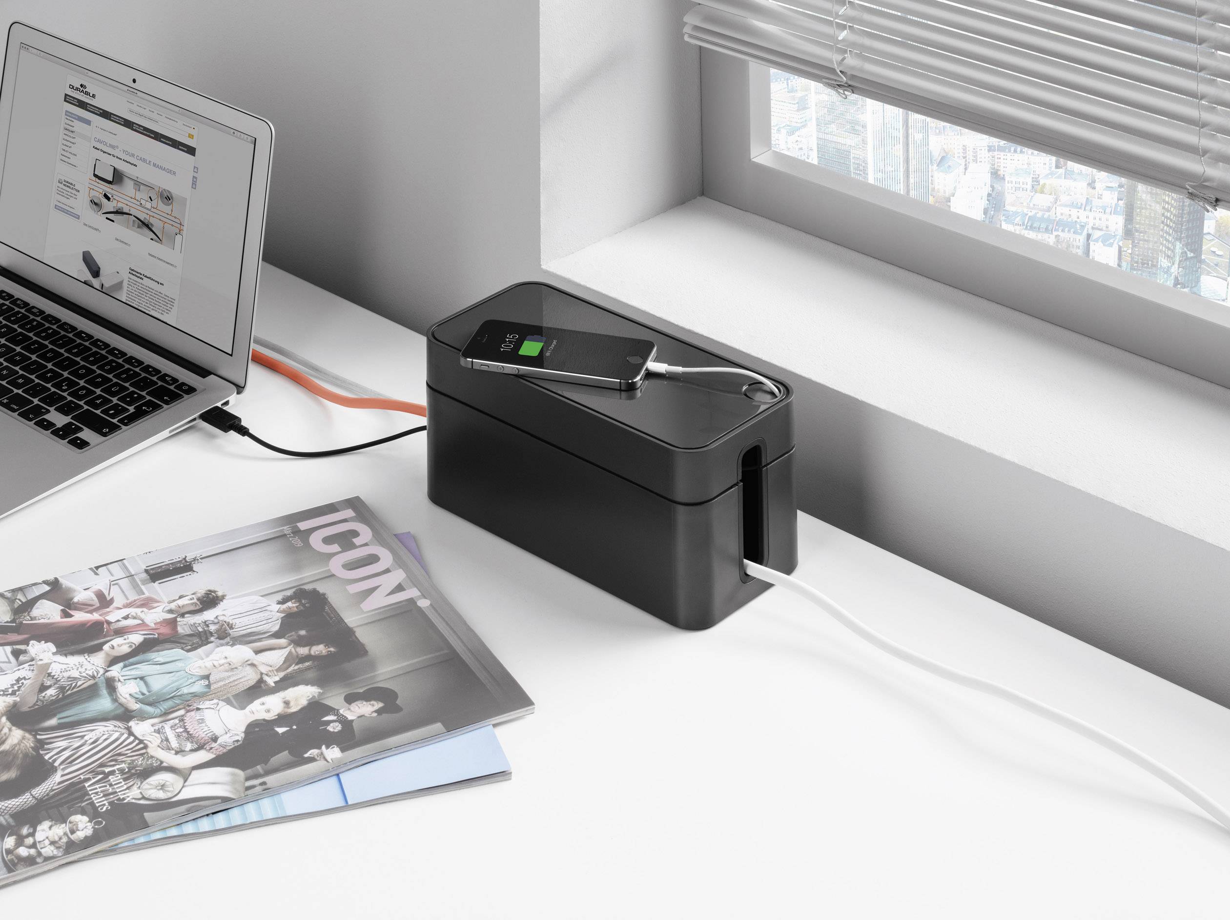 Egy laptop egy kábelvezető dobozhoz csatlakozik. Egy okostelefon tölt a dobozon. A doboz mellett egy magazin fekszik.