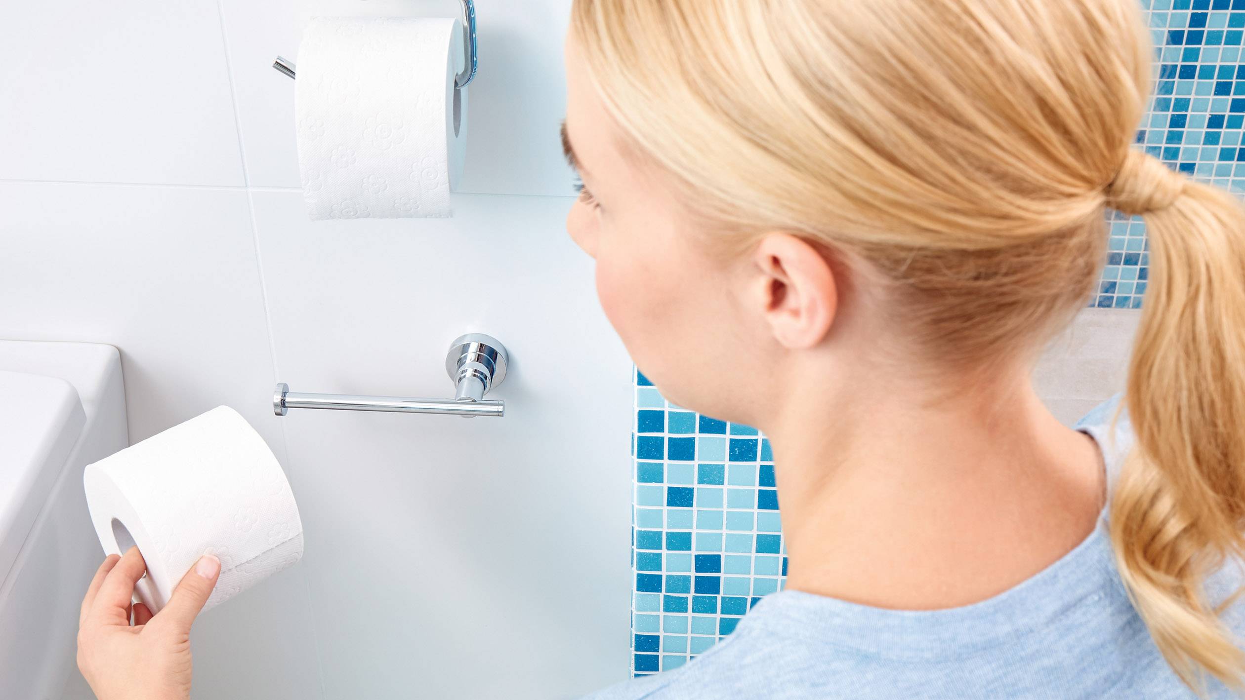 Egy személy a fürdőszobában egy WC-papírtekercs kezében néz egy fali tartóra, amelyen egy másik tekercs van.