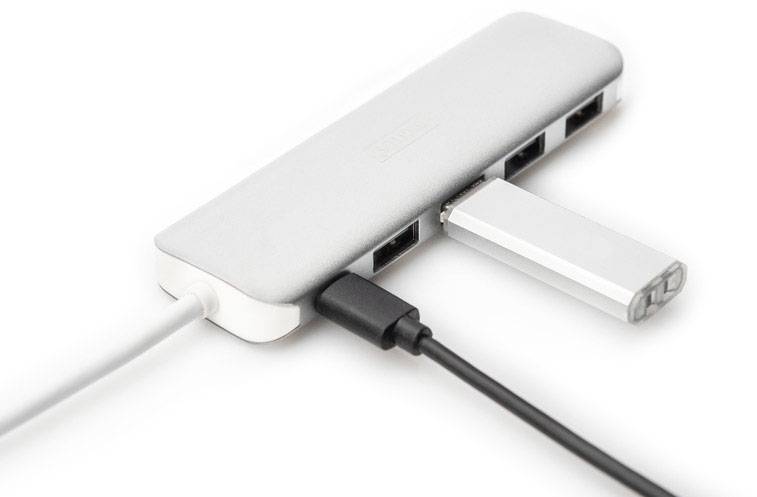 Egy ezüstszínű USB-elosztó több csatlakozóval, amelyhez két kábel és egy pendrive van csatlakoztatva, fehér felületen fekszik.