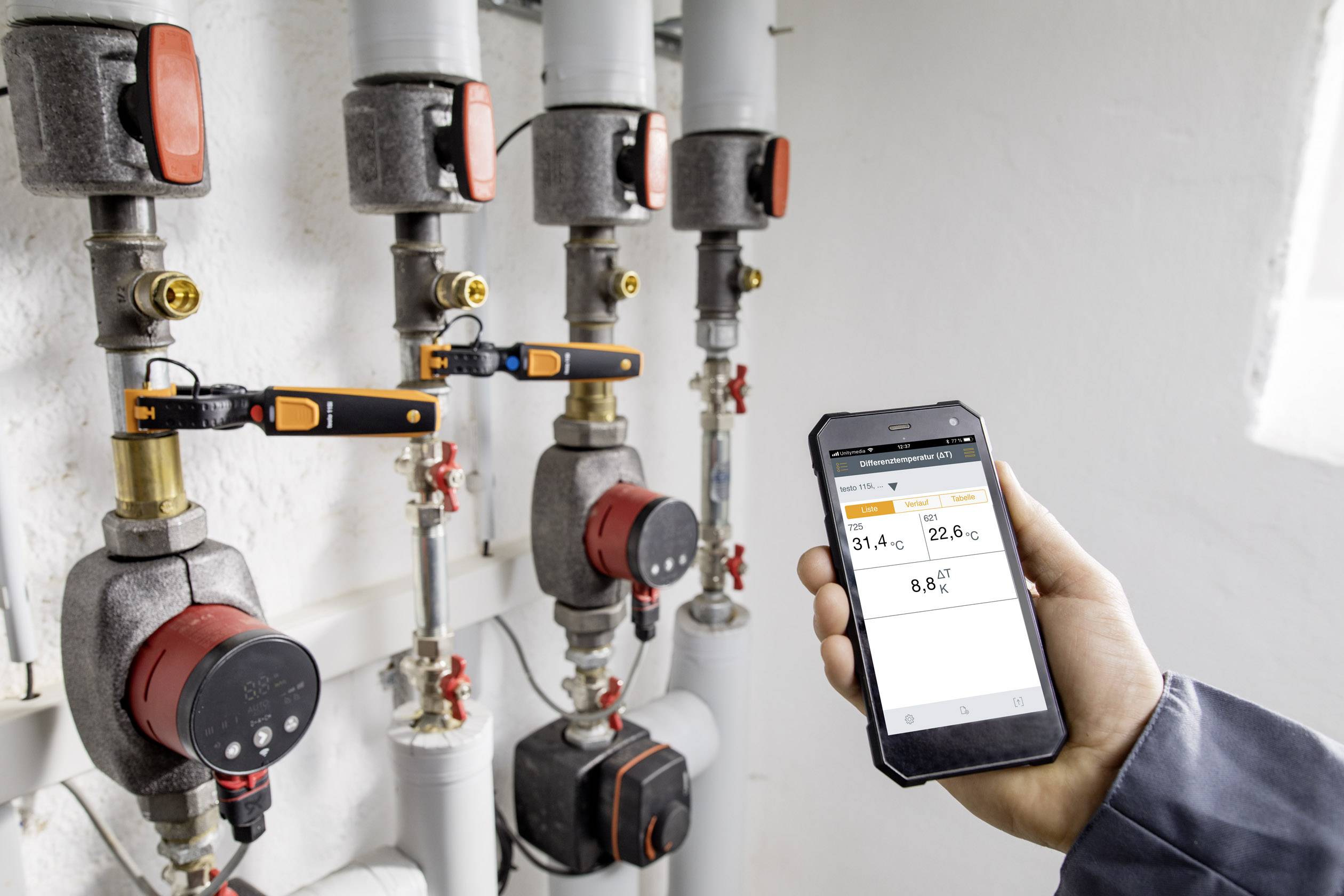 Egy személy egy okostelefont tart, amelyen egy fűtésszabályzó alkalmazás látható. A háttérben több csővezeték szelep és érzékelő figyelhető meg.