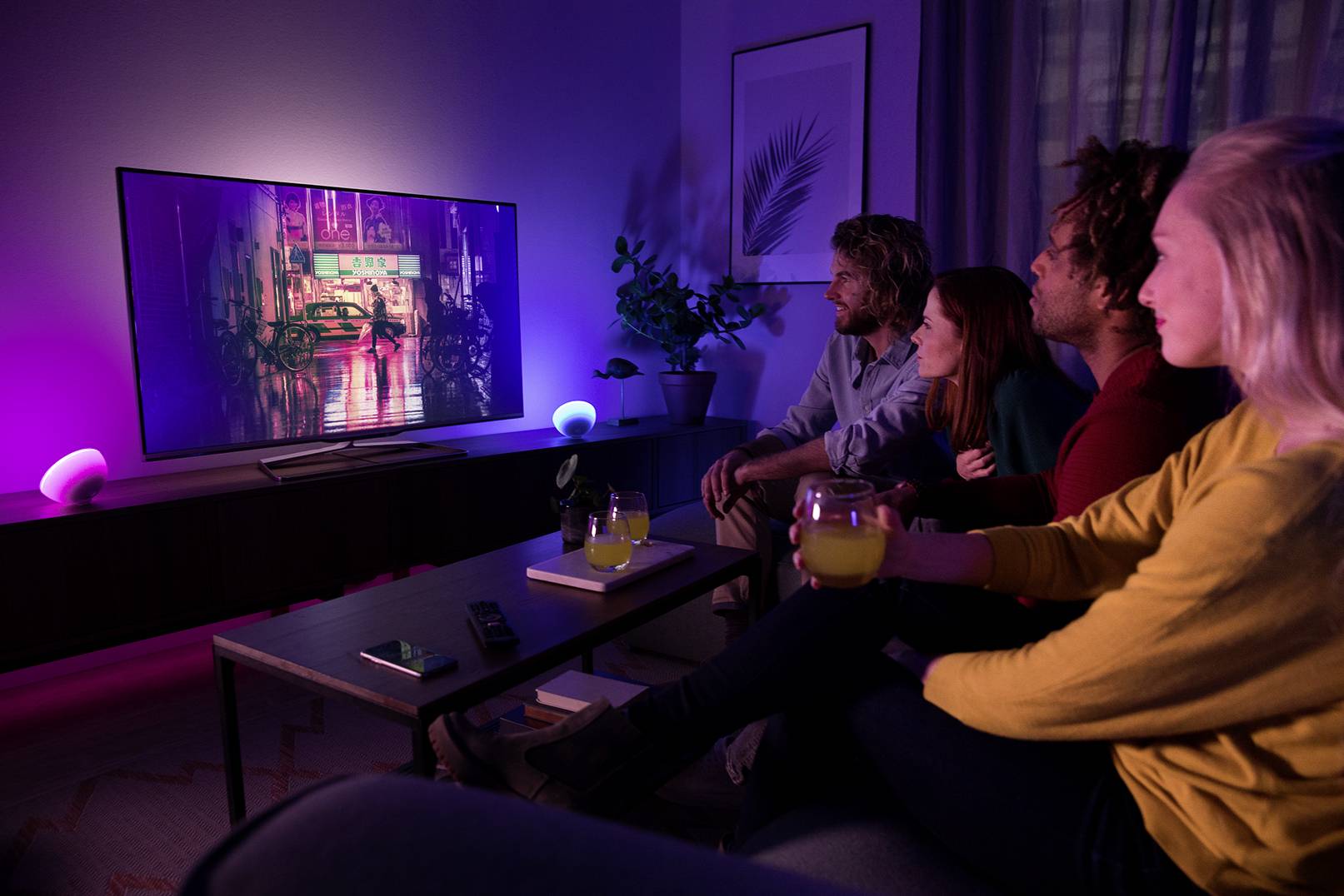 Négy ember ül egy sötétített nappaliban, és egy nagy televízióra néz, amelynek színes háttérvilágítása van.