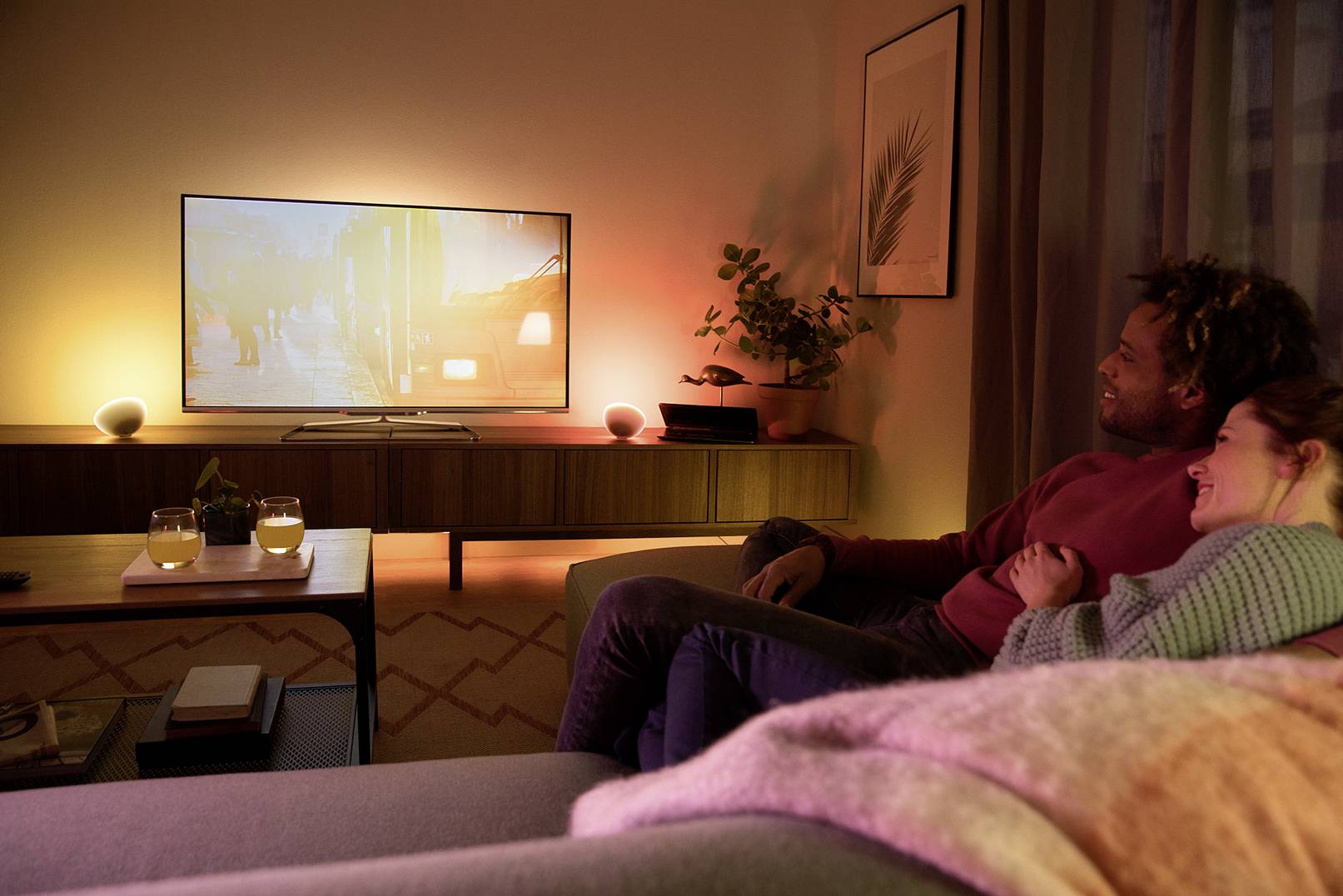 Egy pár kényelmesen ül a kanapén és tévét néz. A szoba meleg fénnyel van megvilágítva. Az asztalnál két ital áll.