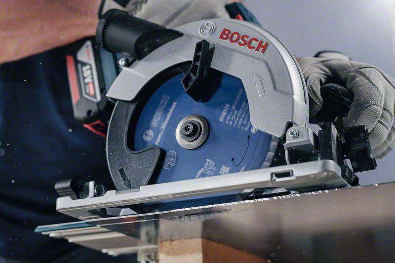Egy személy egy Bosch kézi körfűrésszel faanyagot vág egy műhelyben. A fűrésznek kék fűrészlapja van, és biztonságosan irányítják.