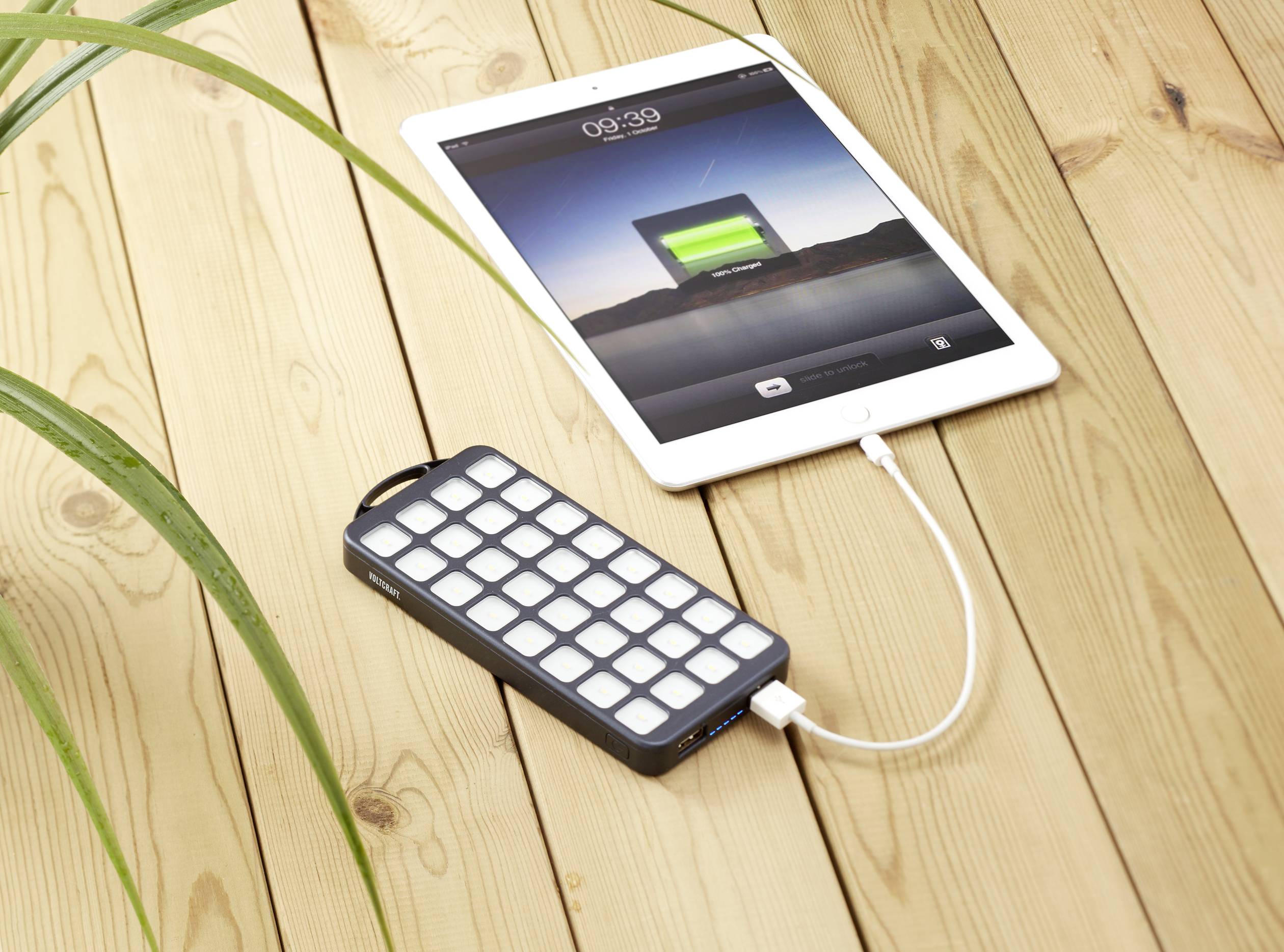 Egy tablet egy fa asztalon egy hordozható powerbank segítségével tölt USB-kábelen keresztül. Egy növény részben látható.