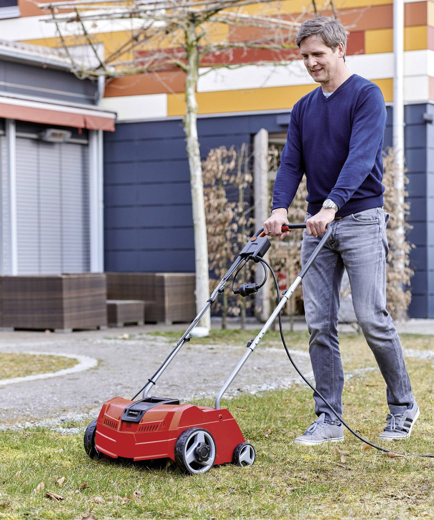 Egy férfi egy piros elektromos fűnyíróval nyírja a füvet egy kertben. A háttérben egy modern épület látható.