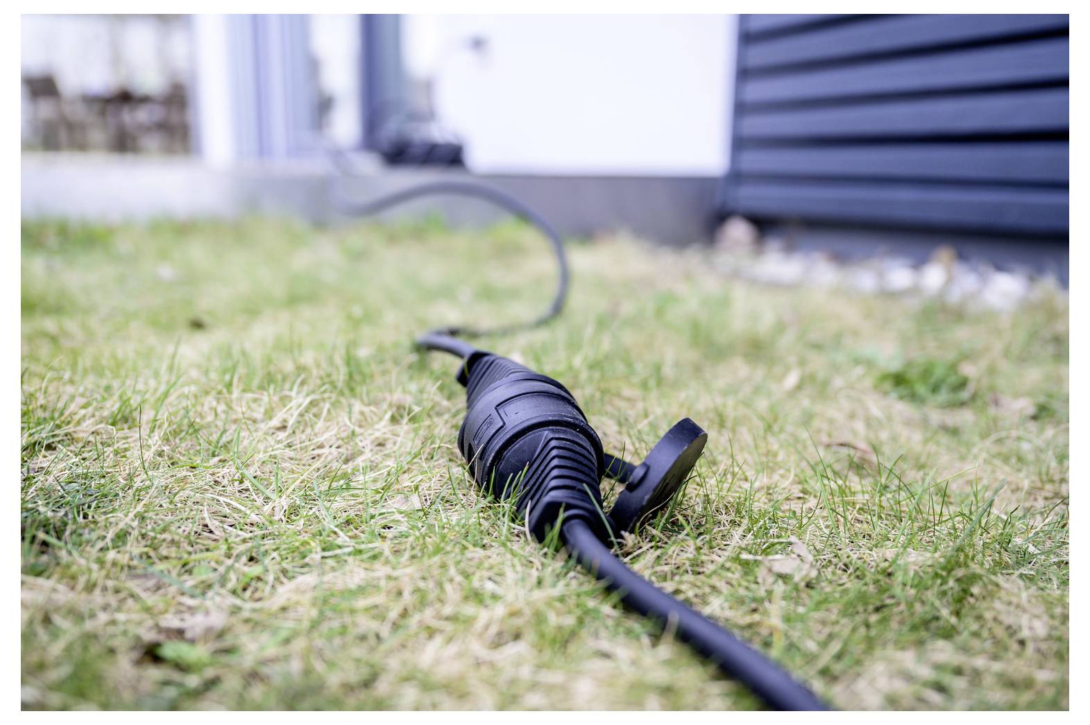 Fekete kültéri elektromos kábel közeli képe, védődugóborítóval a száraz fű mellett a házfal és a burkolat közelében.