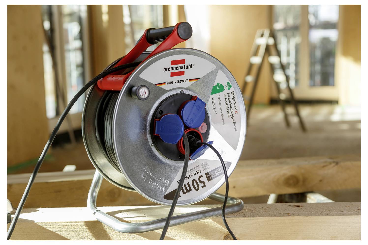 Egy fém kábeldob, amelyen két kék aljzat van, egy fadeszkán helyezkedik el egy részben felépített épületben, nagy ablakokkal és egy létrával.