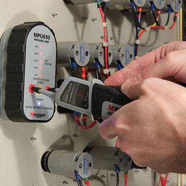 Egy személy digitális mérőműszert használ egy elektromos vezérlőegység feszültségének ellenőrzéséhez. Több kábel van csatlakoztatva.