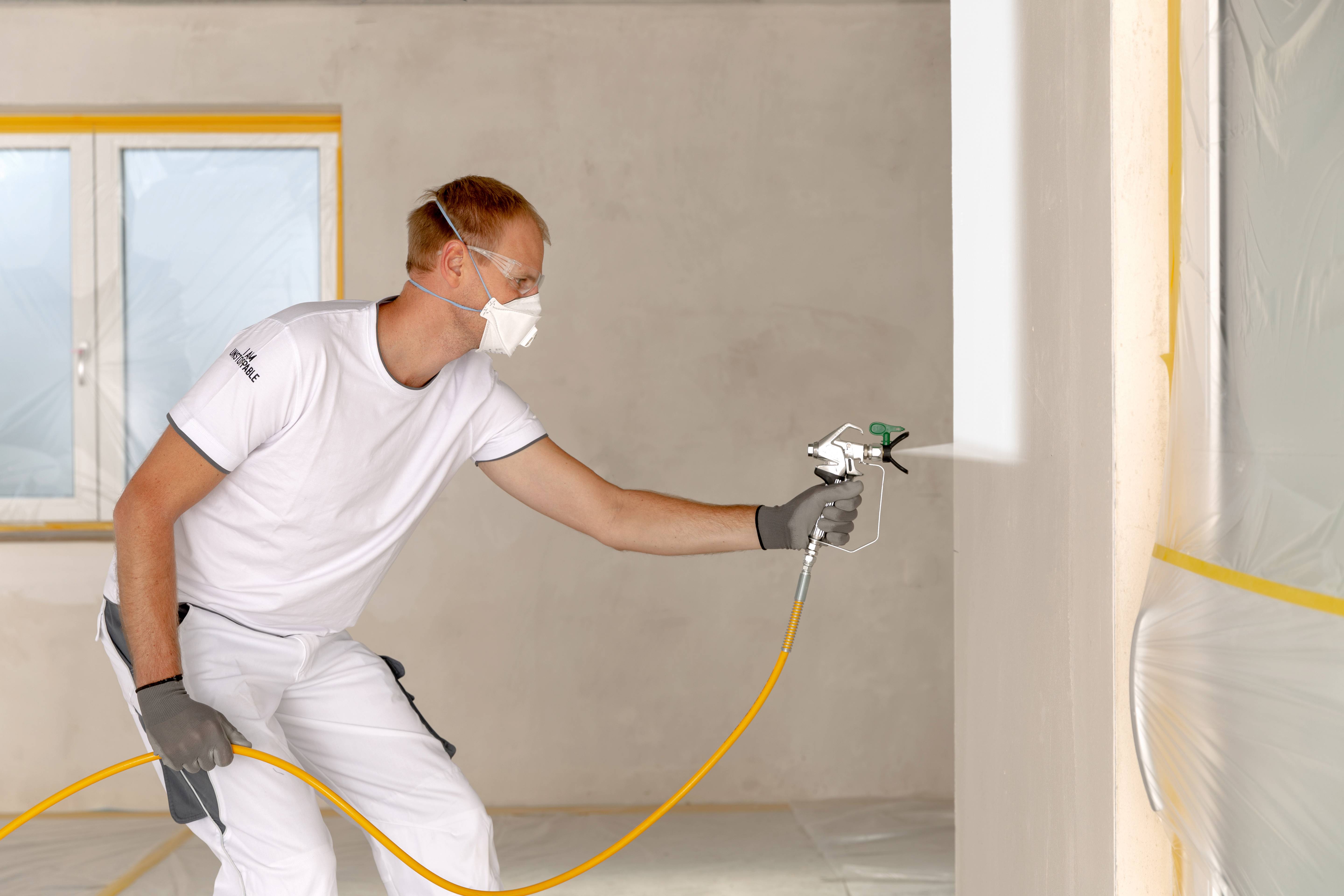 Egy férfi festékszórópisztollyal festéket permetez egy falra egy helyiségben. Védőruhát és maszkot visel.